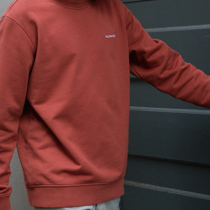 Person, die ein locker sitzendes, rostrotes Sweatshirt und eine helle Hose trägt, steht neben einer dunkelgrauen Wand, einen Arm leicht ausgestreckt, Gesicht nicht sichtbar.