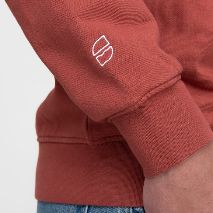 Nahaufnahme des Arms einer Person, die ein rostfarbenes Sweatshirt mit einem kleinen weißen gestickten Logo auf dem Ärmel oberhalb der Manschette trägt. Die Person trägt außerdem eine blaue Jeans.