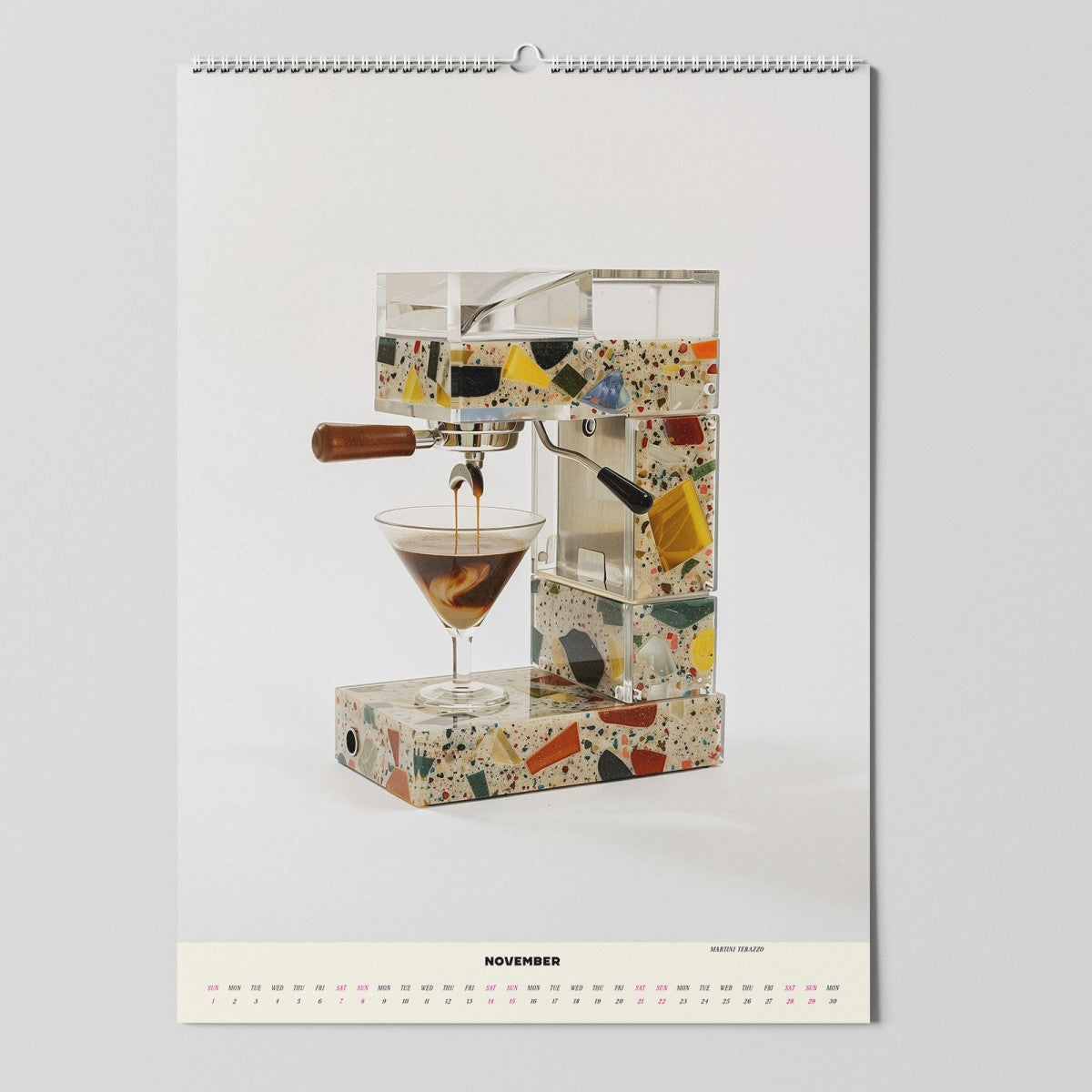 Eine moderne Espressomaschine mit einem farbenfrohen Design im Terrazzo-Stil gießt den Espresso in ein Martiniglas. Das Bild erscheint auf einem Wandkalender, der für den Monat November geöffnet ist.