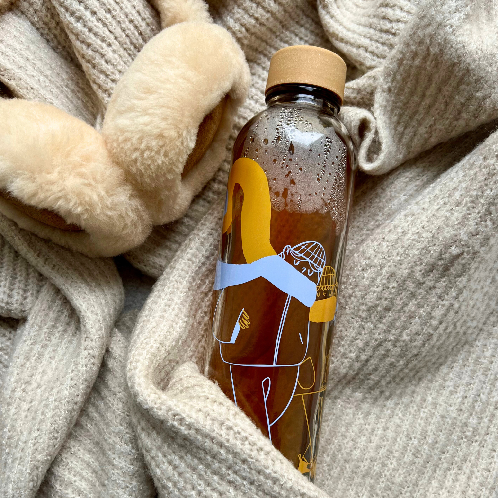Eine Wasserflasche mit abstraktem Design liegt auf einem beigen Wollschal neben flauschigen Ohrenschützern.