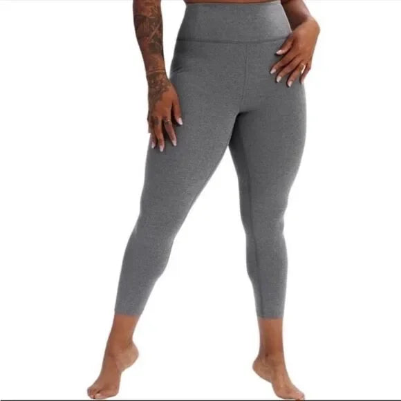 Eine Person mit Tätowierungen auf dem linken Arm trägt eine knöchellange, graue Leggings mit hoher Taille. Sie ist barfuß und steht vor einem weißen Hintergrund. Ihr Kopf ist auf dem Bild nicht zu sehen.