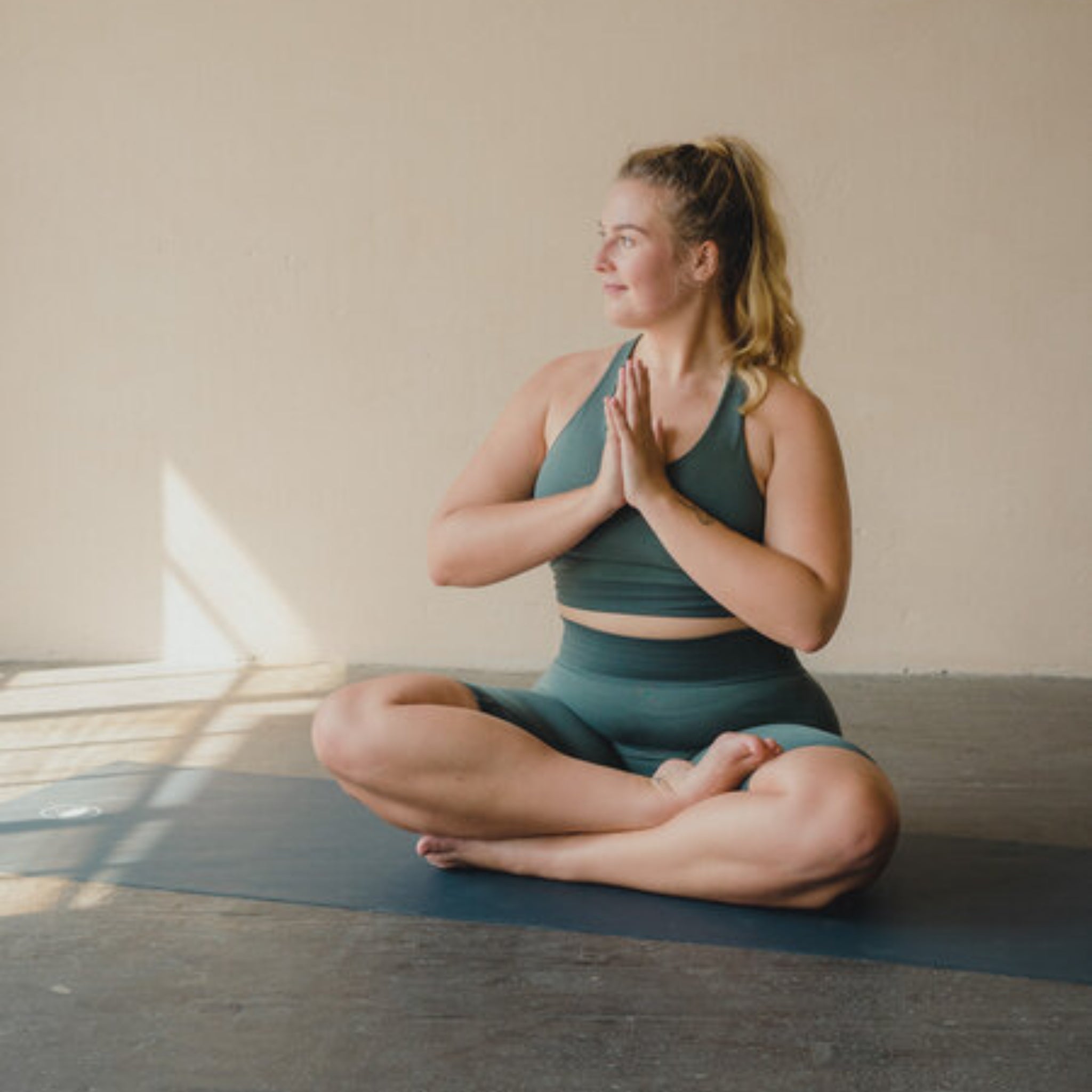 Frau in grüner Sportkleidung sitzt im Lotussitz auf einer Yogamatte, Hände in Gebetshaltung, schaut zur Seite.