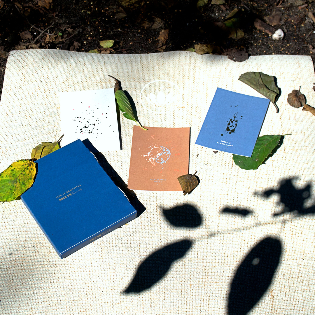 Blaues Notizbuch und drei Karten auf beigem Stoff, umgeben von Blättern und Schatten im Sonnenlicht.