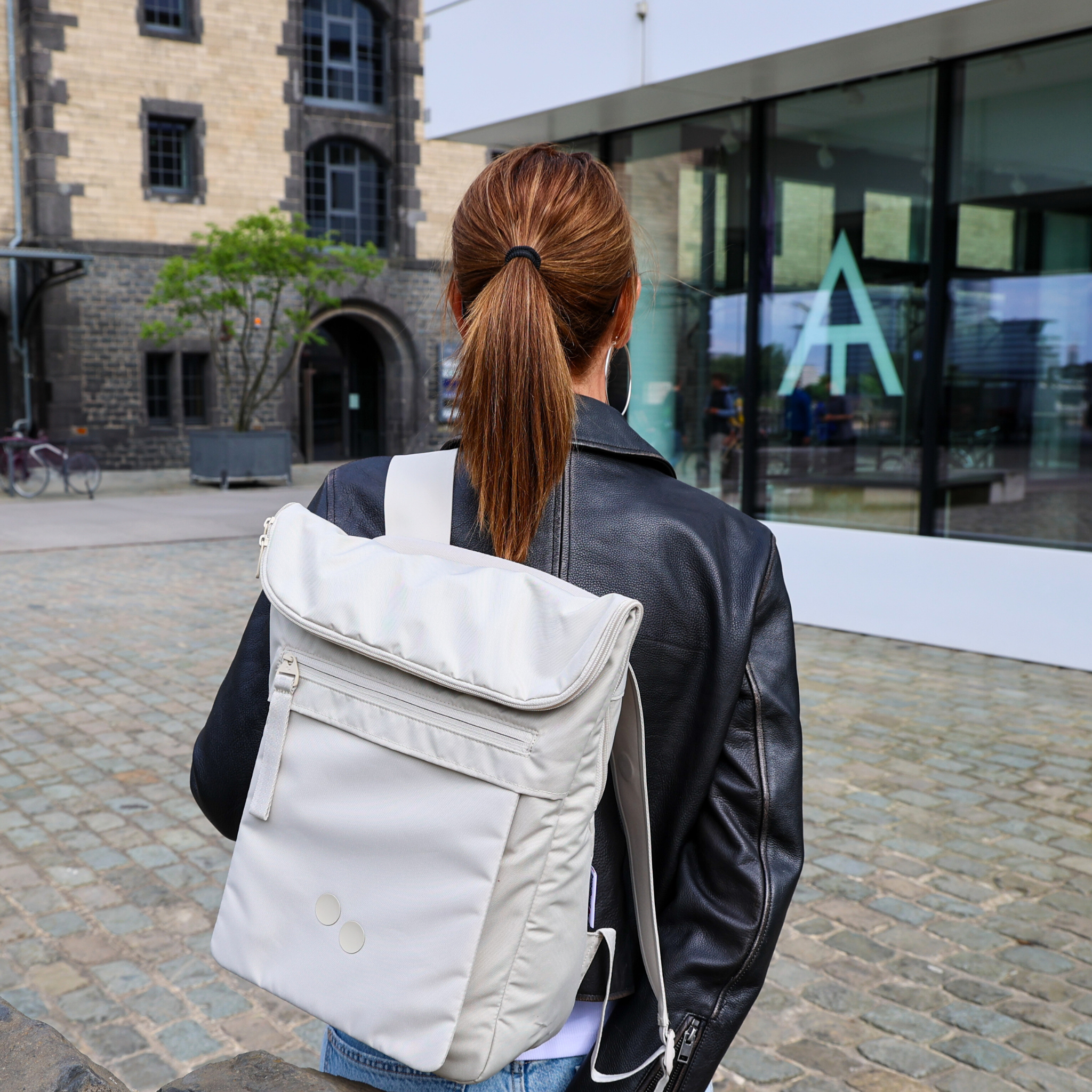 Frau mit weißem Rucksack und Lederjacke steht vor modernen und historischen Gebäuden auf einem gepflasterten Platz.