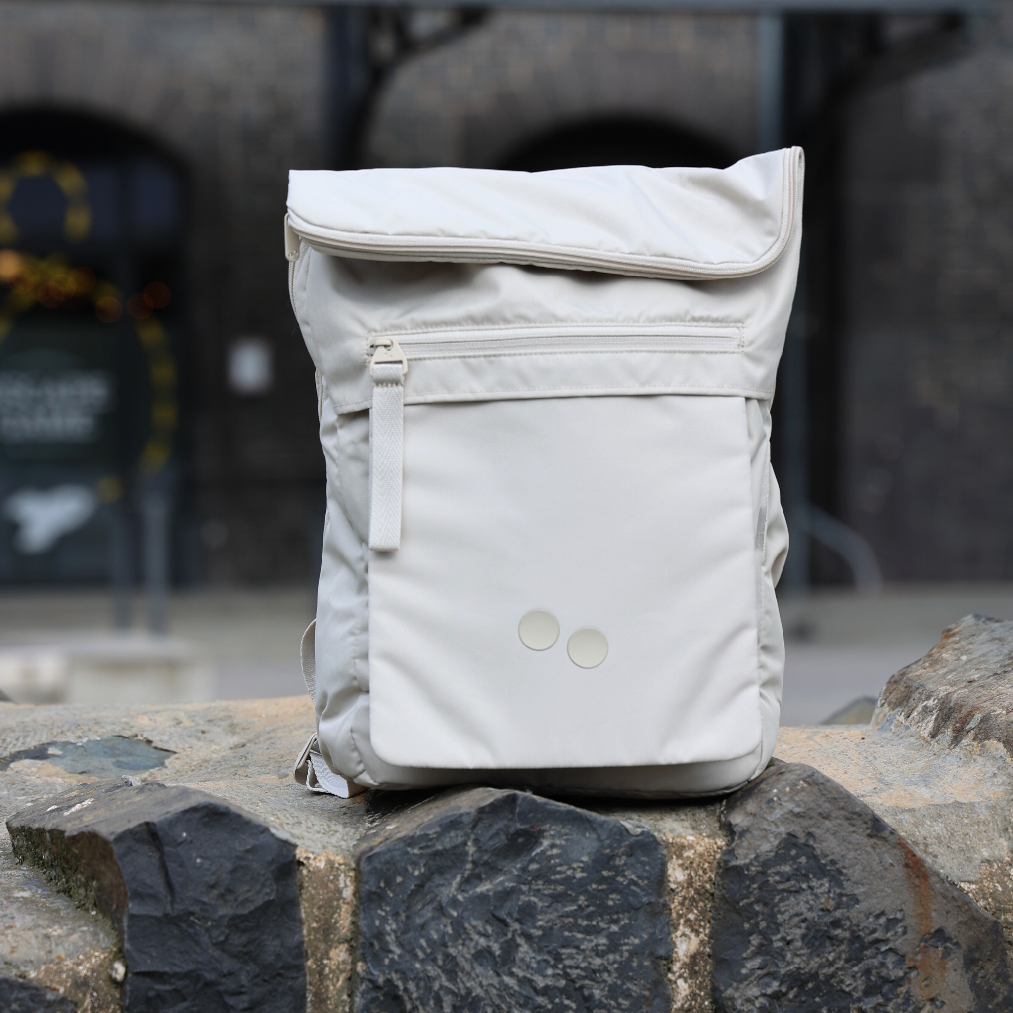 Beiger Rucksack steht auf grobem Steinuntergrund mit unscharfem, dunklem Gebäude im Hintergrund.