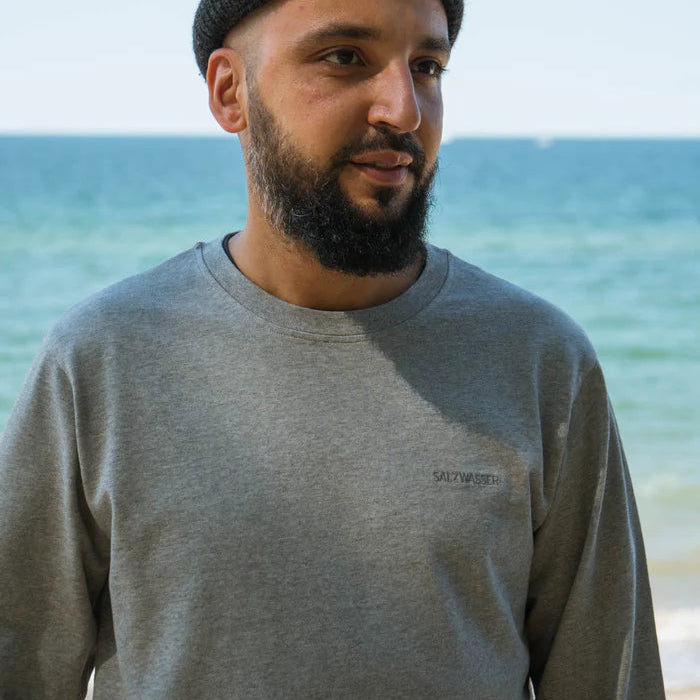 Ein Mann mit Bart, grauem Sweatshirt und dunkler Mütze steht am Meer, mit blauem Wasser und Himmel im Hintergrund.
