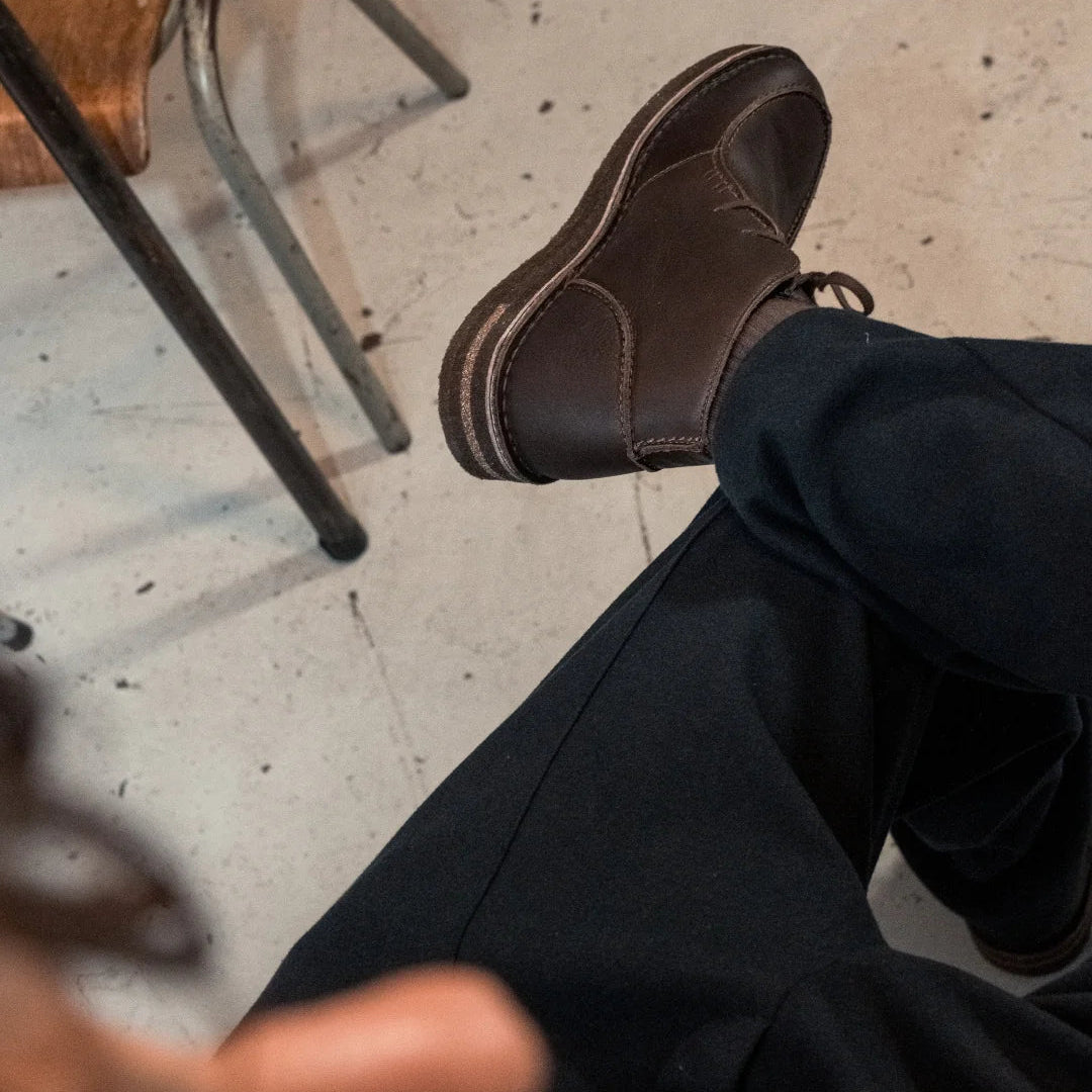 Eine mit einer dunklen Hose bekleidete Person sitzt mit gekreuzten Beinen auf einem hellen Boden. Nur ihr Unterkörper und ihre braunen Lederschuhe sind sichtbar. In der Nähe steht ein Holzstuhl mit Metallbeinen.