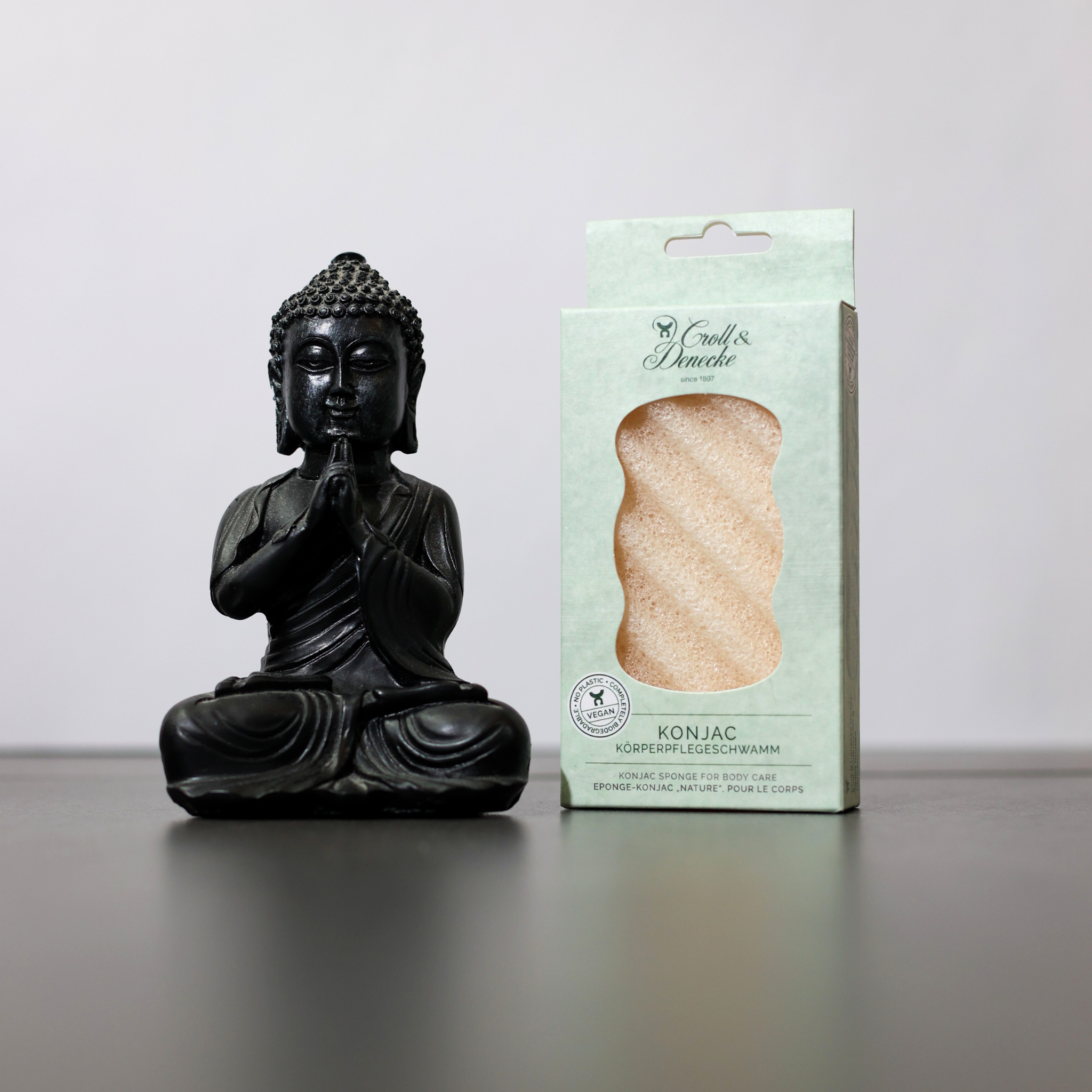 Schwarze Buddha-Statue neben einer Verpackung eines Konjac-Körperschwamms auf einem grauen Hintergrund.