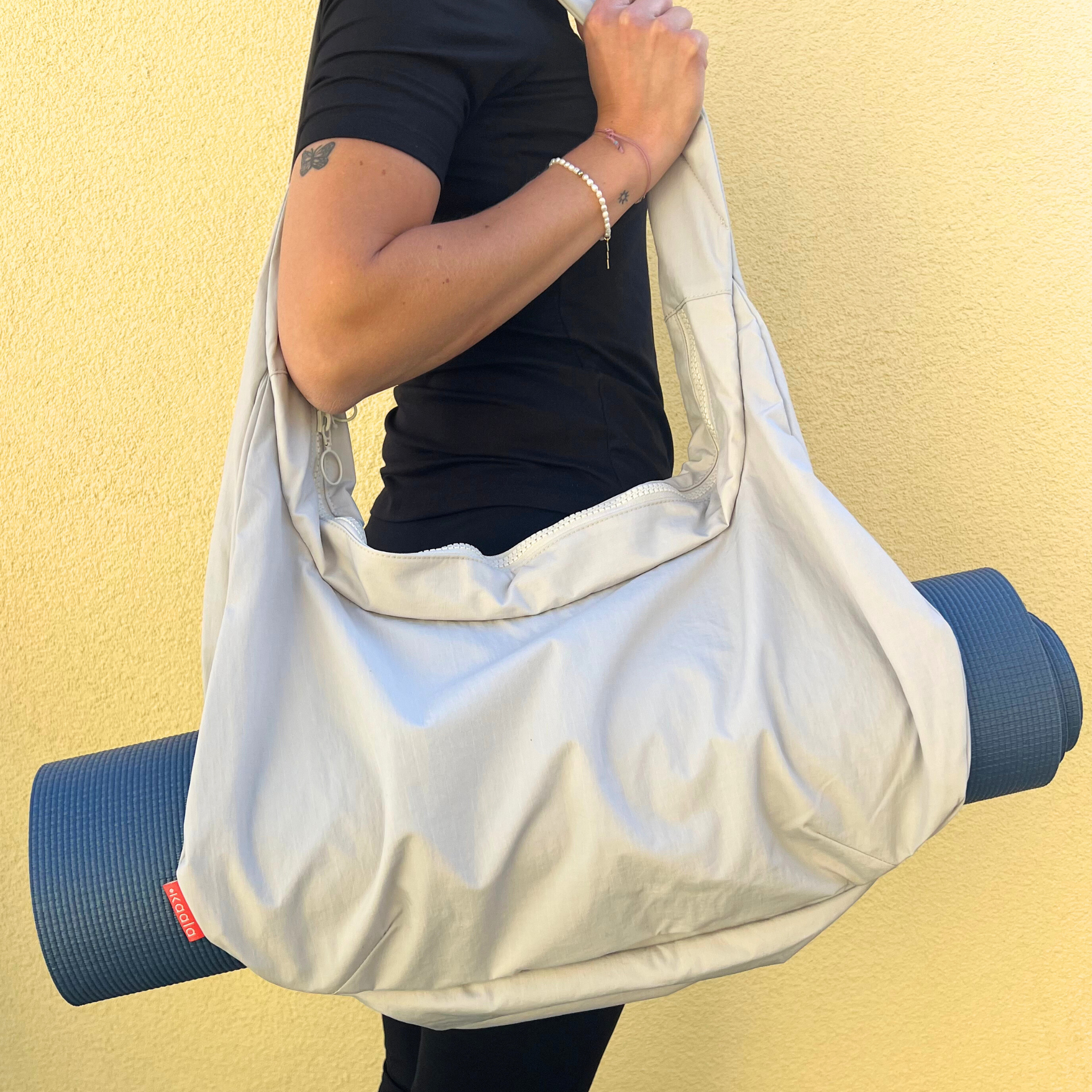 Person trägt eine große, helle Tasche mit einer blauen Yogamatte darin vor einem gelben Hintergrund.