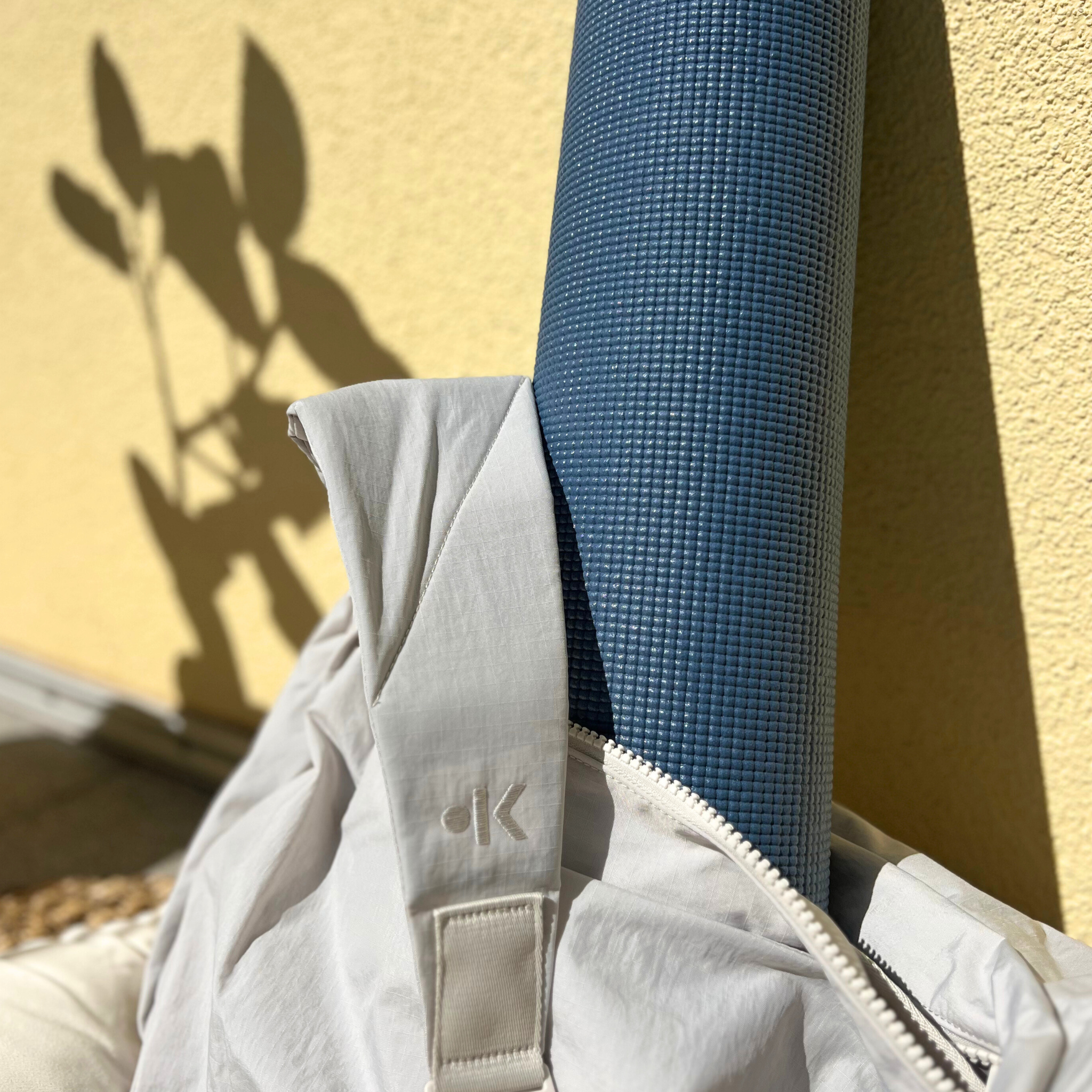 Graue Sporttasche mit blauer Yogamatte, die gegen eine gelbe Wand lehnt, mit Pflanzen im Schatten auf der Wand.