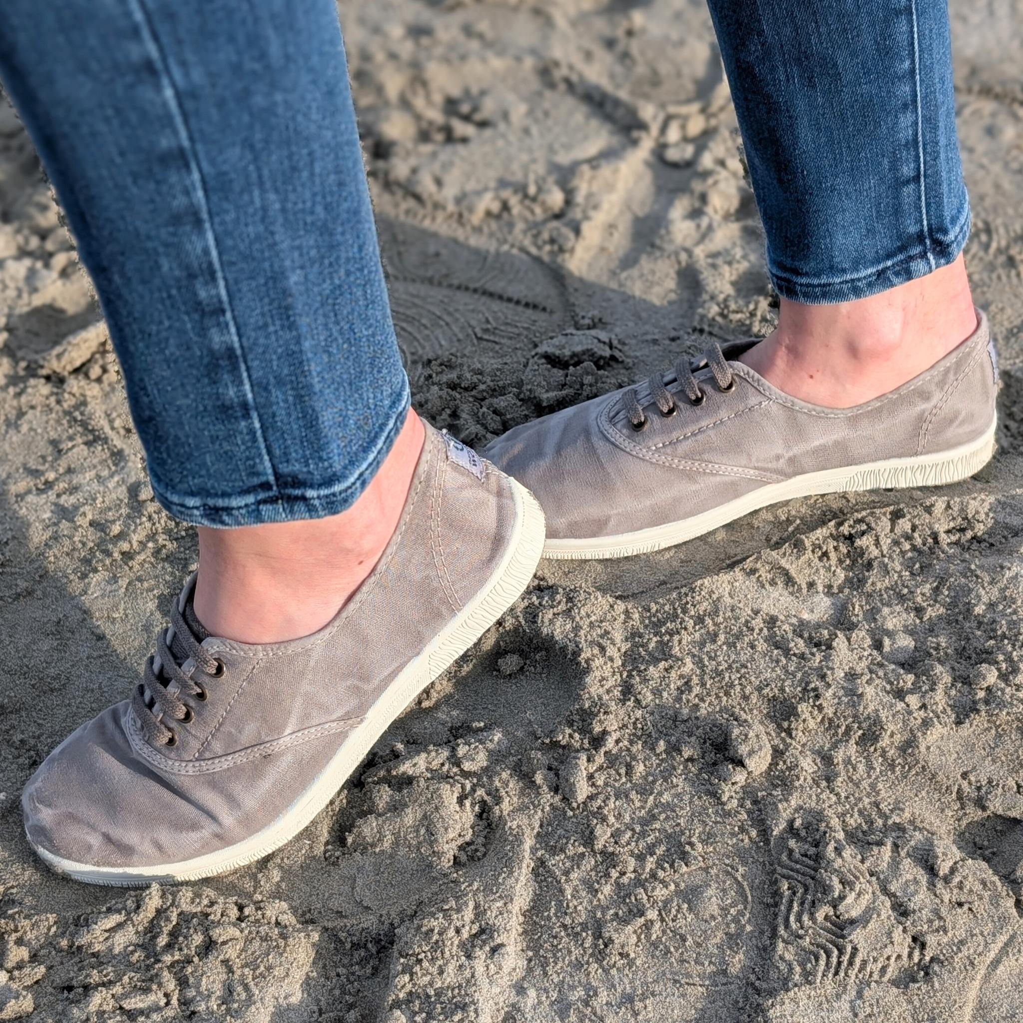 Person in blauen Jeans und grauen Turnschuhen steht auf sandigem Boden.