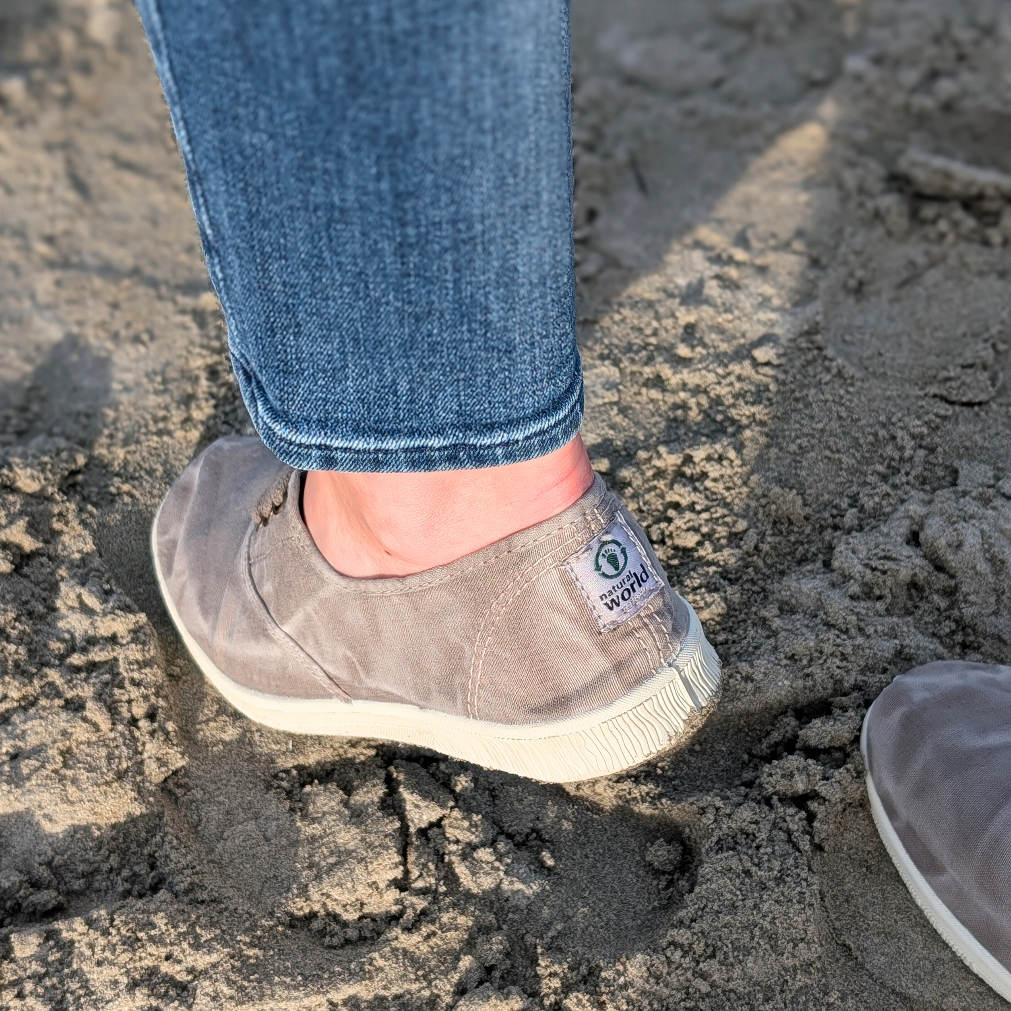 Eine Person trägt graue Stoffschuhe und blaue Jeans, stehend auf sandigem Boden.