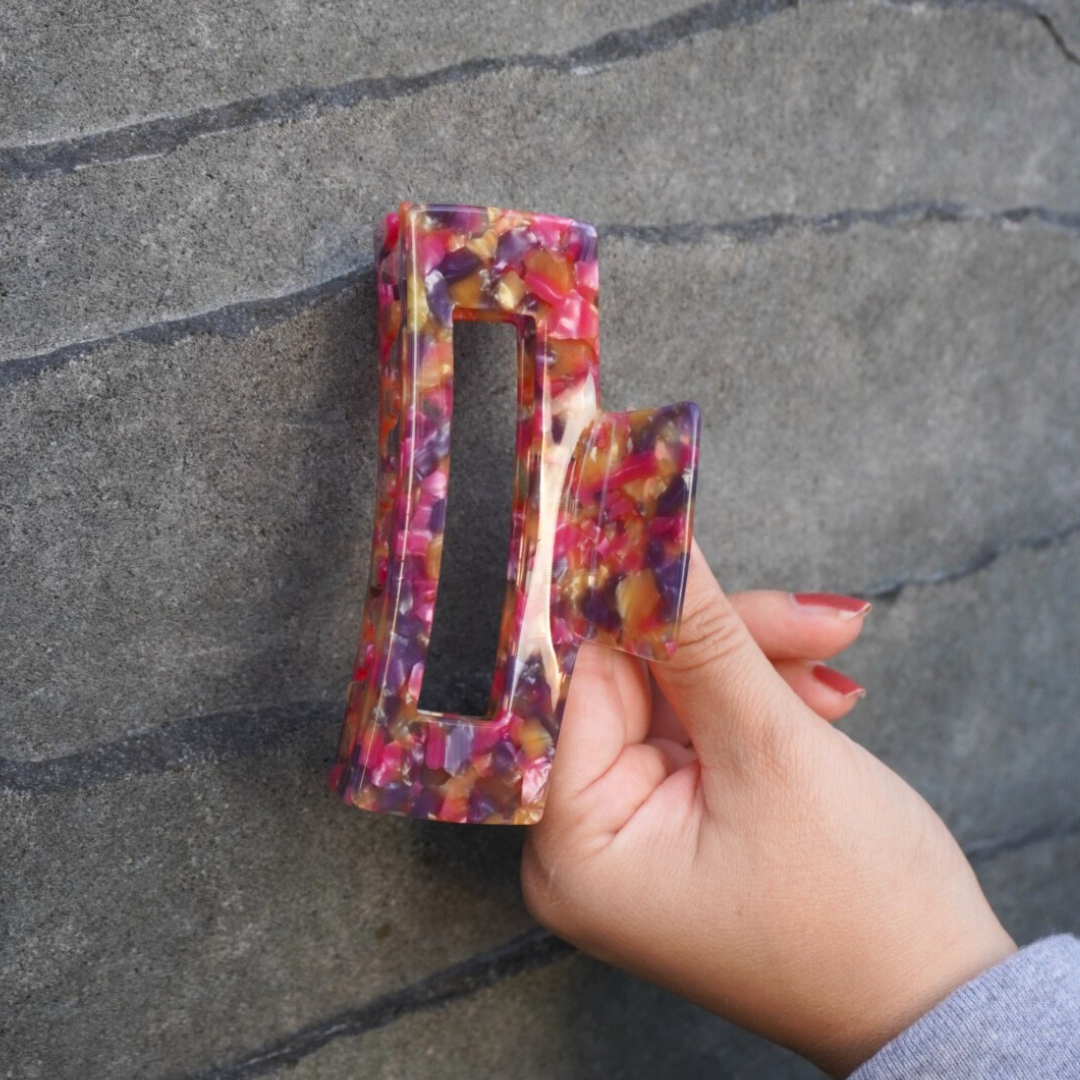 Hand hält bunte Haarspange aus Kunststoff vor einer grauen Steinwand.