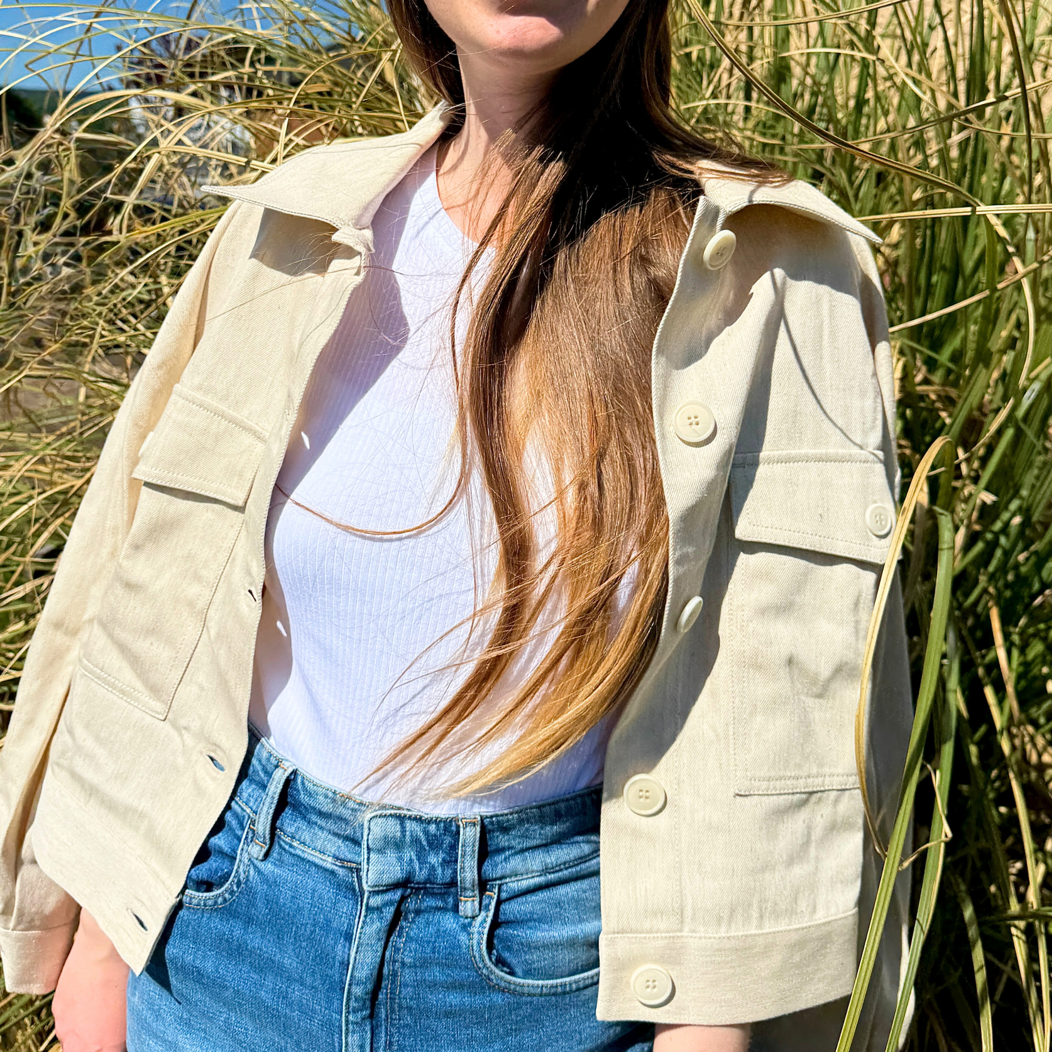 Frau in weißem T-Shirt und beigener Jacke steht vor hohen Gräsern, ihr Haar weht leicht im Wind.
