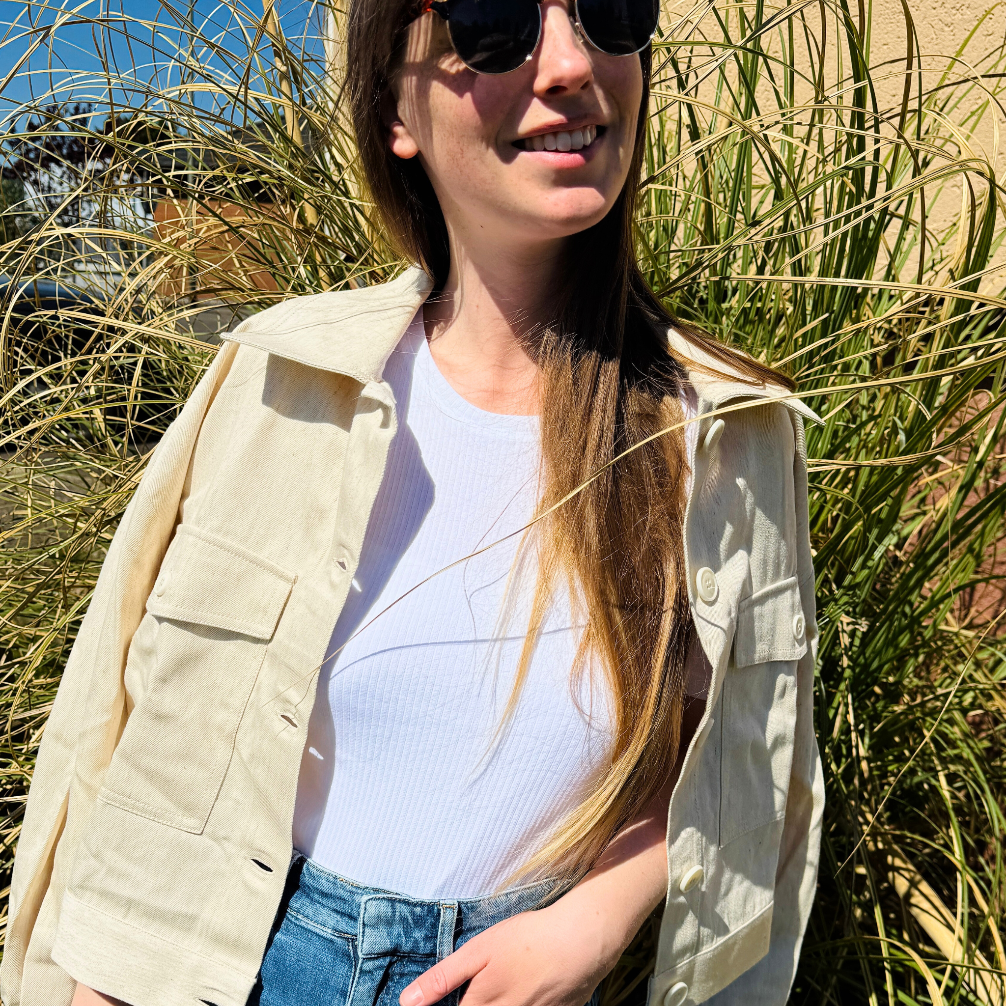 Frau mit Sonnenbrille und heller Jacke steht lächelnd vor hohen Gräsern in der Sonne.