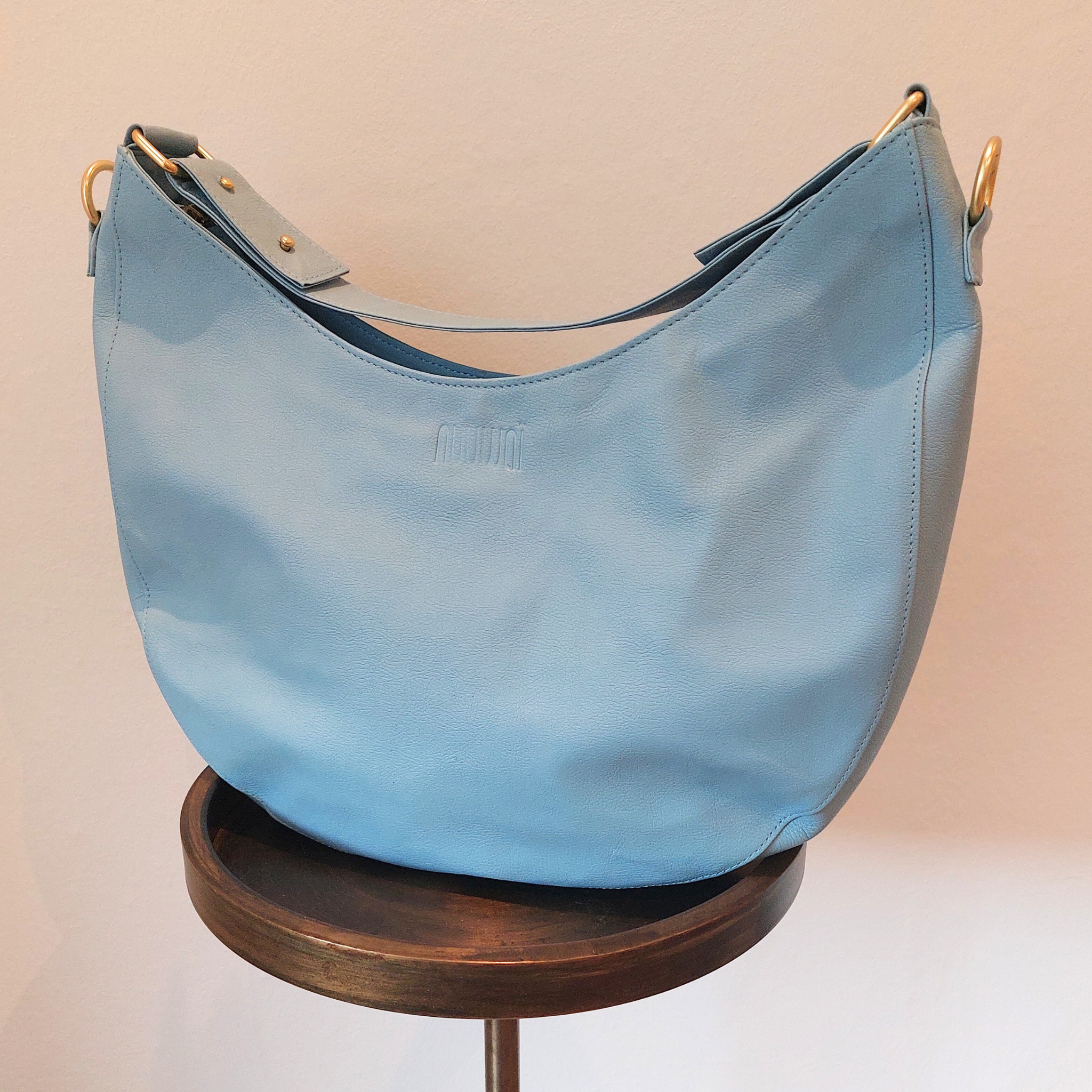 Blaue Ledertasche mit rundem Design, auf einem Holzhocker ausgestellt.