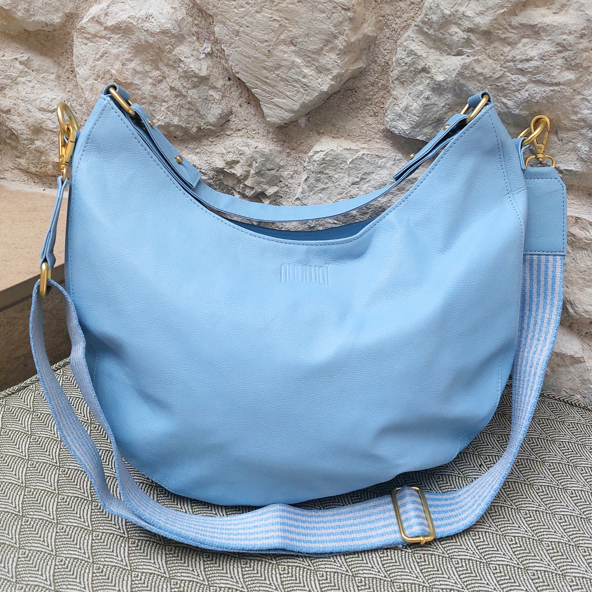 Hellblaue Handtasche mit goldenen Schnallen und gestreiftem Schultergurt vor einer Steinwand.