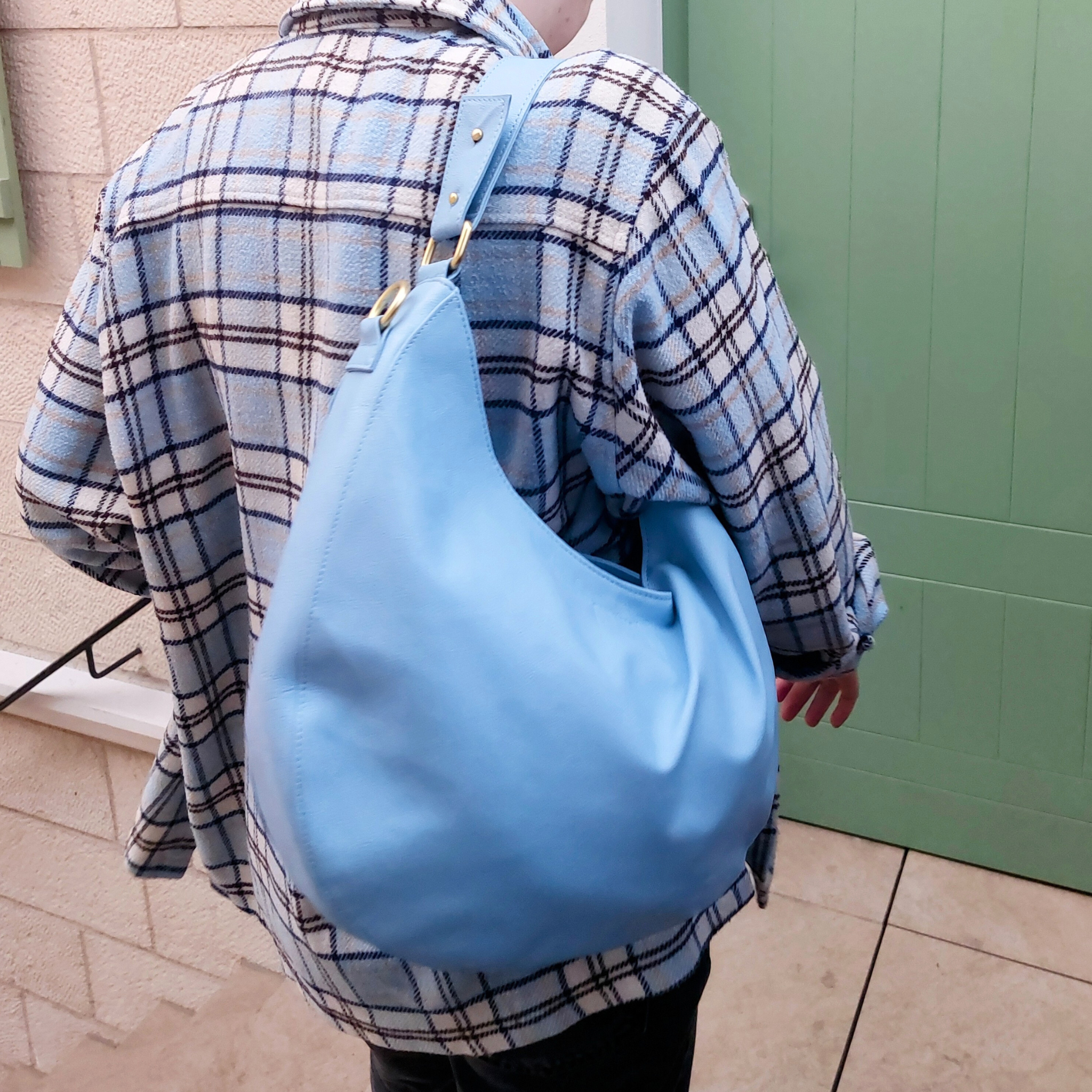 Person in karierter Jacke trägt eine große blaue Tasche über der Schulter und geht in Richtung einer grünen Tür.