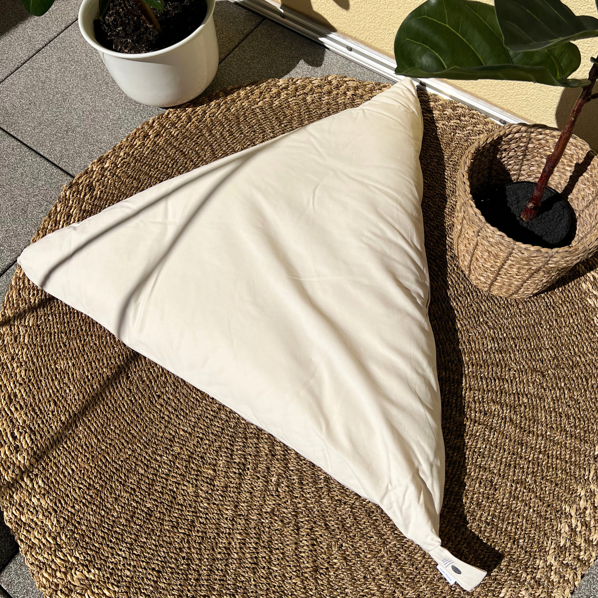Dreieckiges Kissen auf einem geflochtenen Teppich neben zwei Pflanzen in Töpfen auf einer Terrasse.