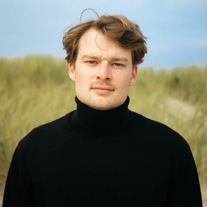Ein junger Mann mit hellbraunem Haar, der einen schwarzen Rollkragenpullover trägt, steht im Freien vor hohem Gras, mit einem bewölkten blauen Himmel im Hintergrund.