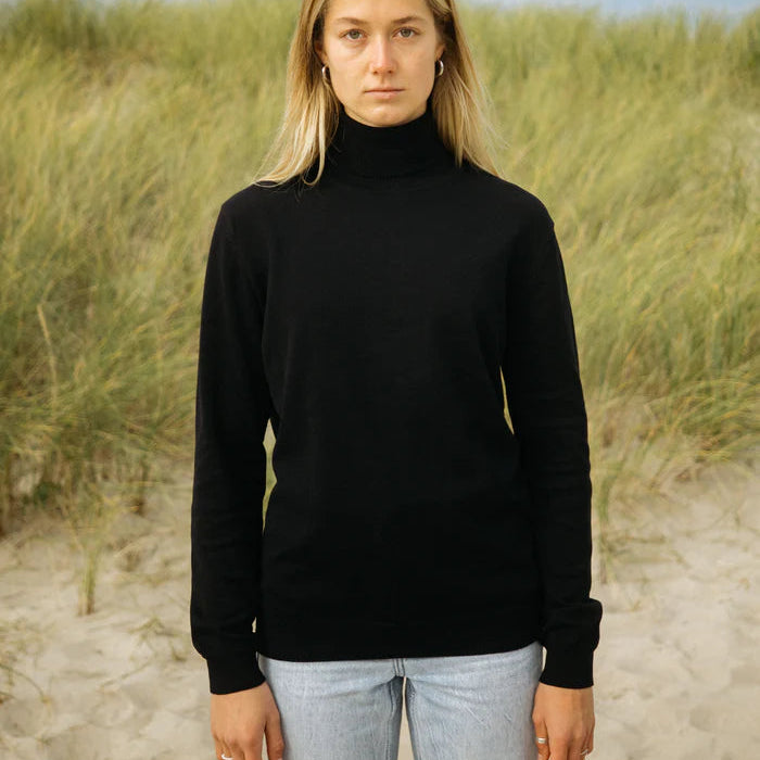 Eine Frau mit blondem Haar, die einen schwarzen Rollkragenpullover und hellblaue Jeans trägt, steht an einem Sandstrand mit hohem Gras im Hintergrund. Sie schaut mit neutralem Blick direkt in die Kamera.