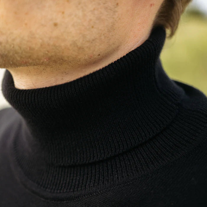 Nahaufnahme einer Person, die einen schwarzen Rollkragenpullover trägt, der einen Teil ihres Kinns, ihres Halses und ihrer Kieferpartie zeigt. Der Hintergrund ist unscharf und im Freien.
