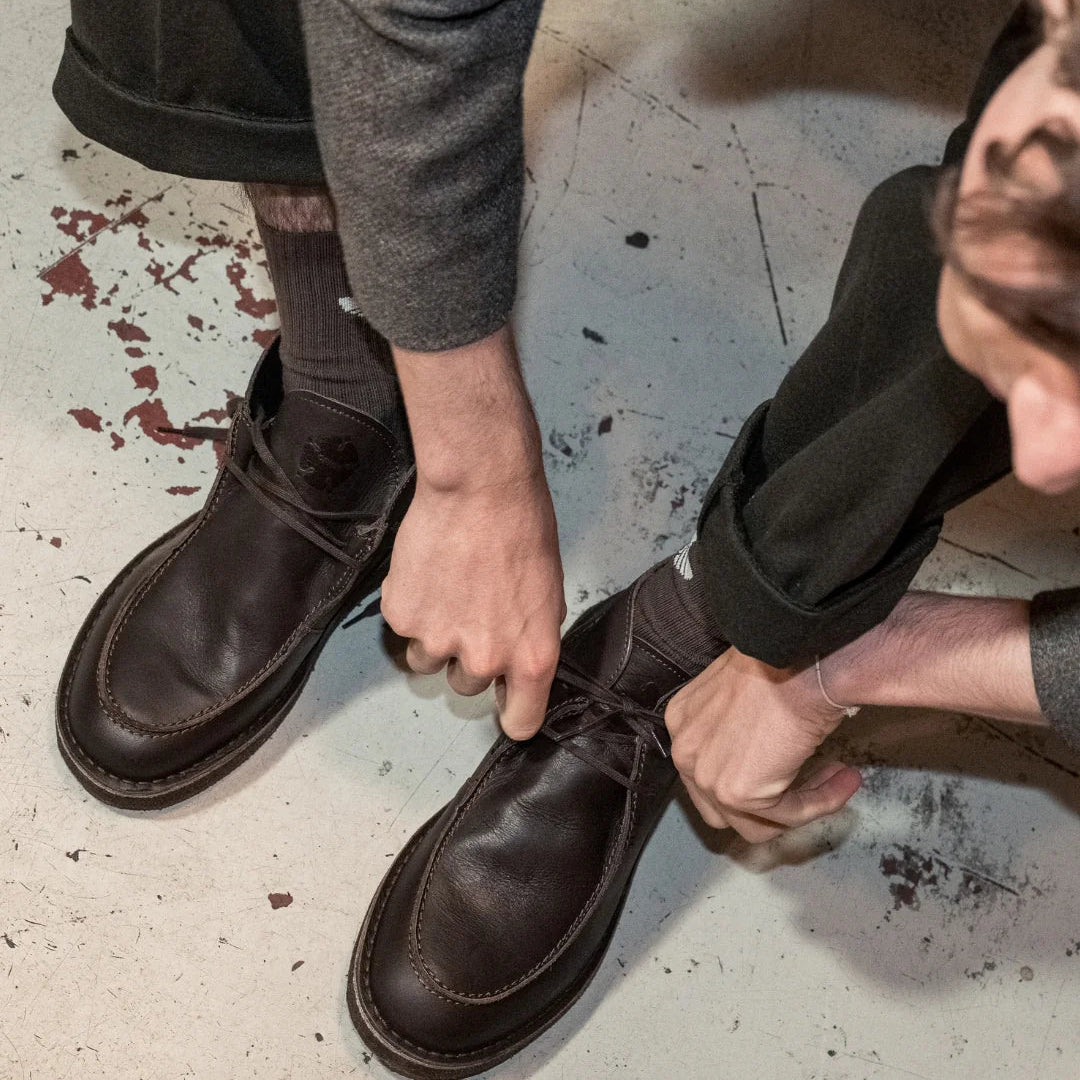 Eine Person in dunkler Hose und grauer Jacke bückt sich, um die Schnürsenkel der dunkelbraunen Lederschuhe zu binden, wobei sie mit einem Fuß leicht nach vorne auf dem abgenutzten, gesprenkelten Boden steht.