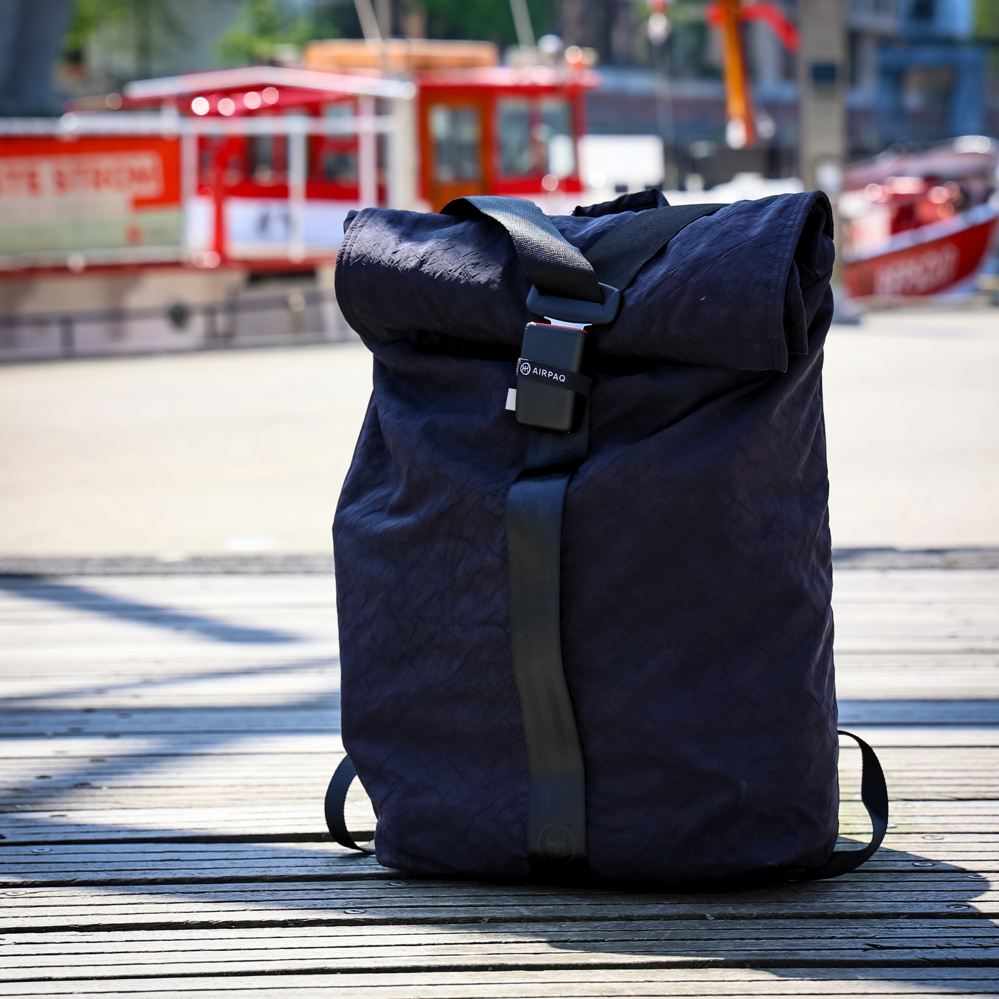 Ein dunkelblauer Rucksack steht auf einem Holzboden, im Hintergrund sind unscharfe rote Boote am Hafen zu sehen.