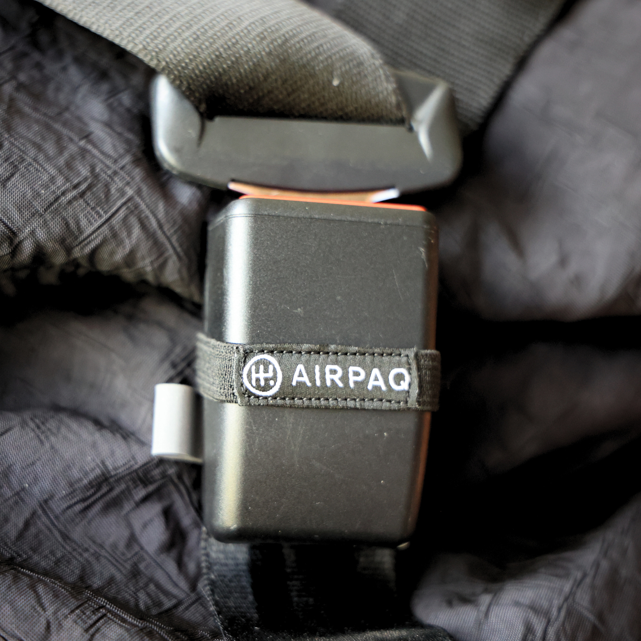 Nahaufnahme eines schwarzen Sicherheitsgurts mit einer Befestigung, auf der "AIRPAQ" steht, auf einem dunklen Stoff.