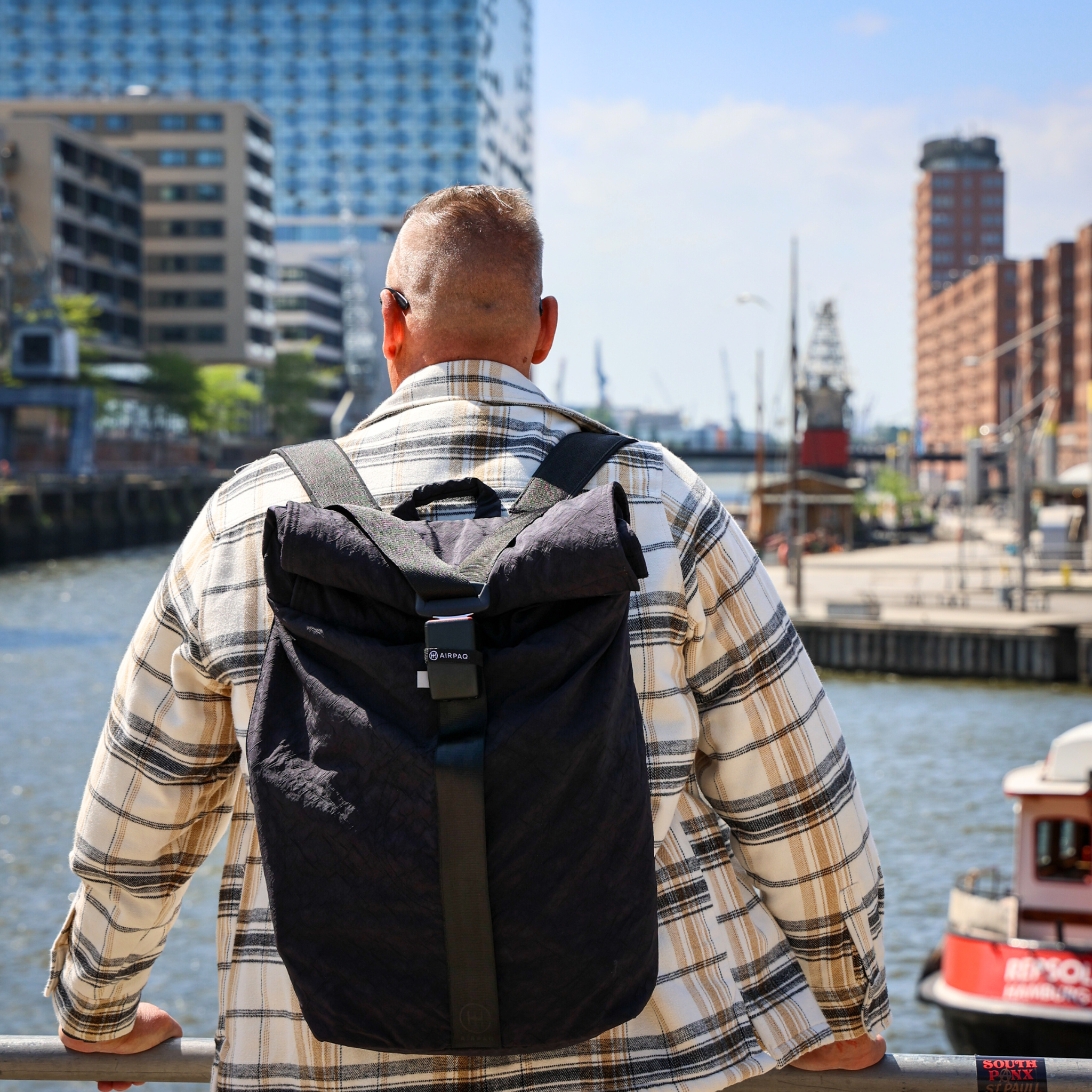 Ein Mann mit Rucksack steht an einem Geländer und blickt auf eine städtische Wasserlandschaft.