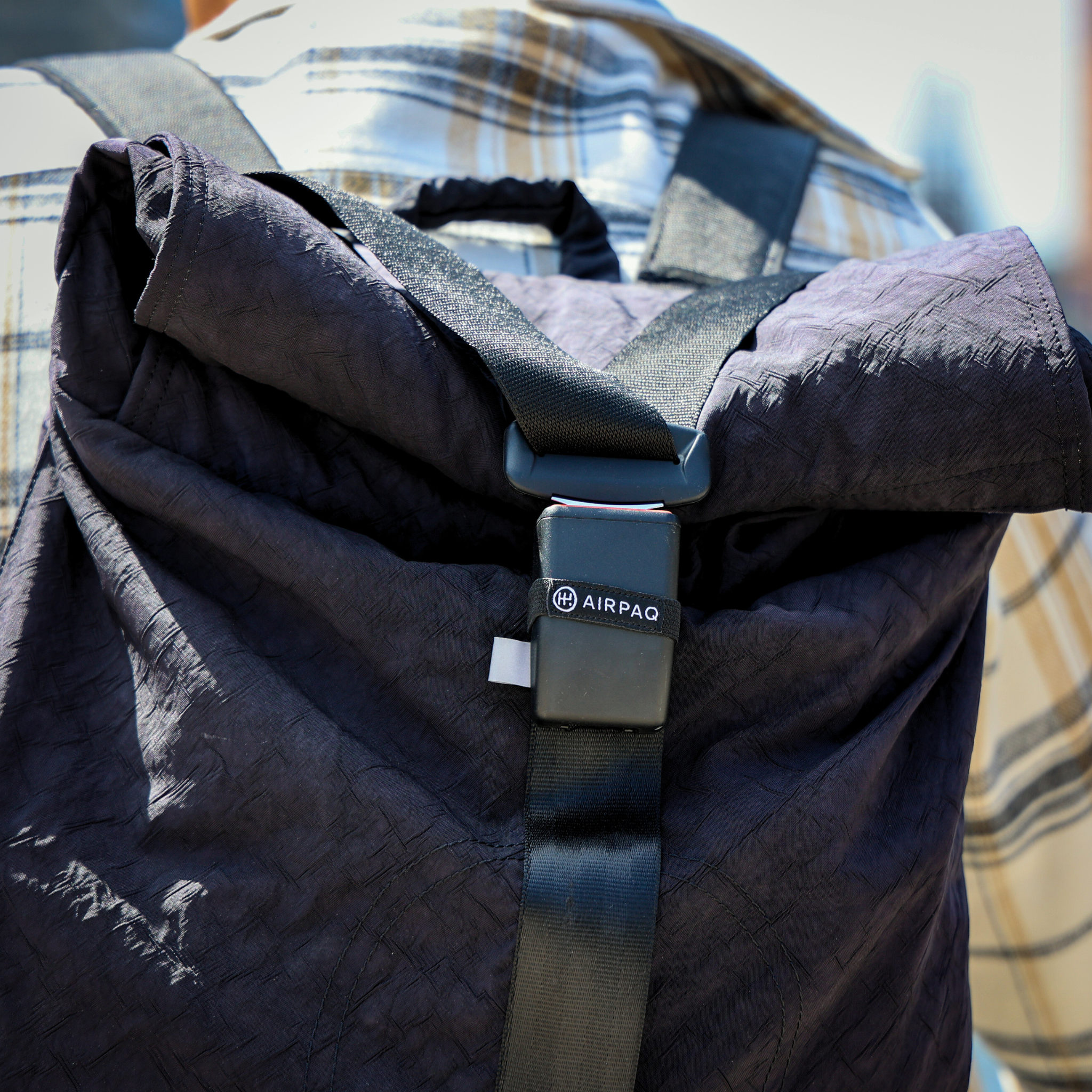 Eine Person trägt einen schwarzen Rucksack mit dem Logo "AIRPAQ" und einer Karosjacke im Hintergrund.