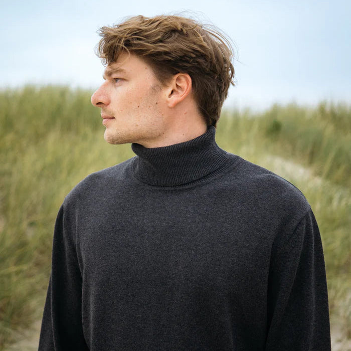 Ein junger Mann mit hellbraunem Haar, der einen dunklen Rollkragenpullover trägt, steht im Freien und blickt zur Seite, während im Hintergrund hohes Gras und ein bewölkter Himmel zu sehen sind.