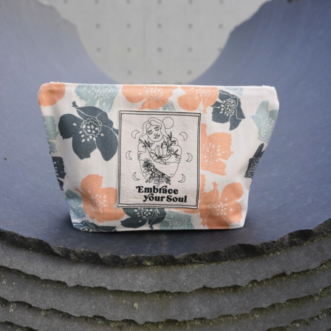 Kosmetiktasche mit floralem Muster und der Aufschrift "Embrace Your Soul", auf einem grauen Untergrund präsentiert.