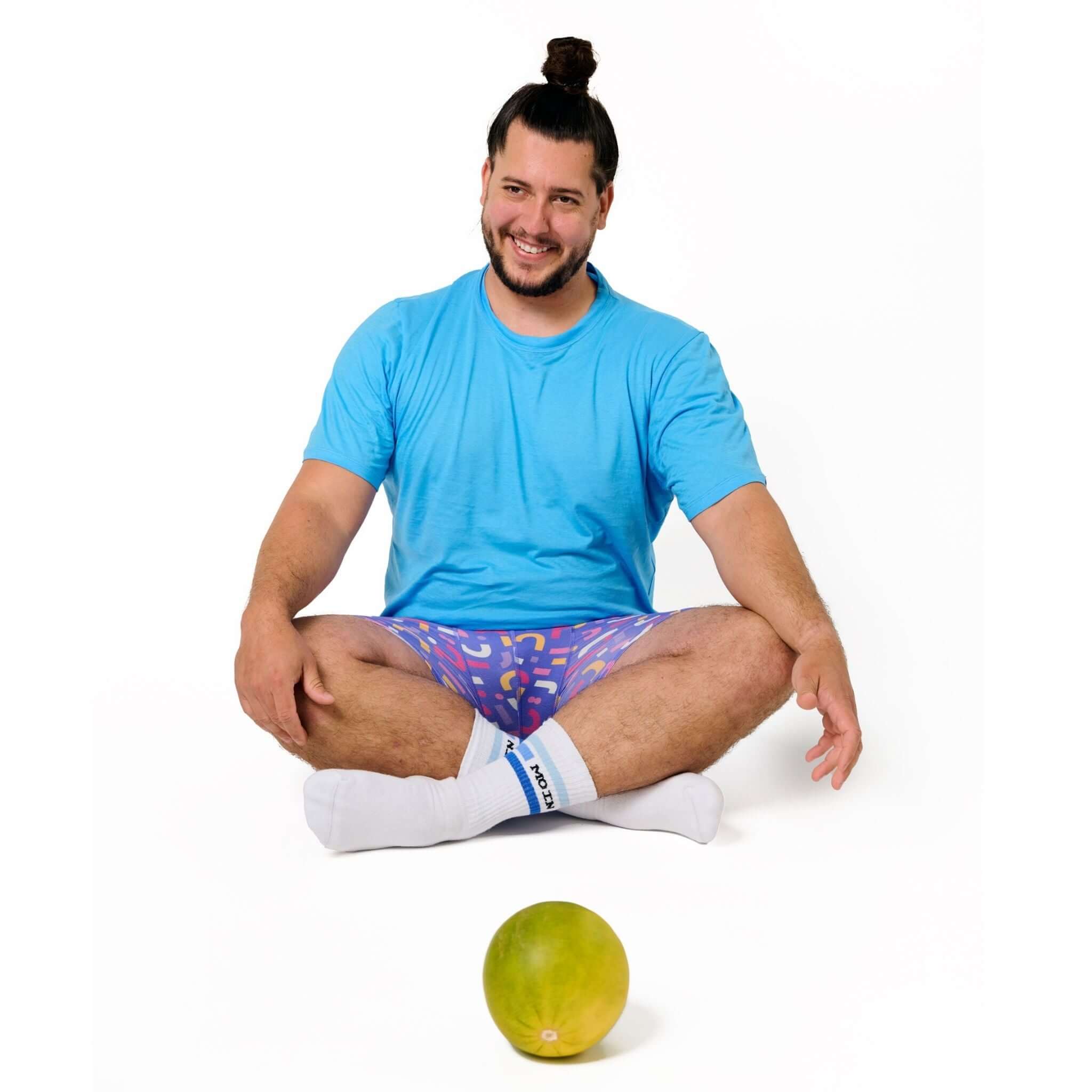 Mann im blauen T-Shirt und bunten Shorts sitzt auf dem Boden mit einer Melone vor sich.