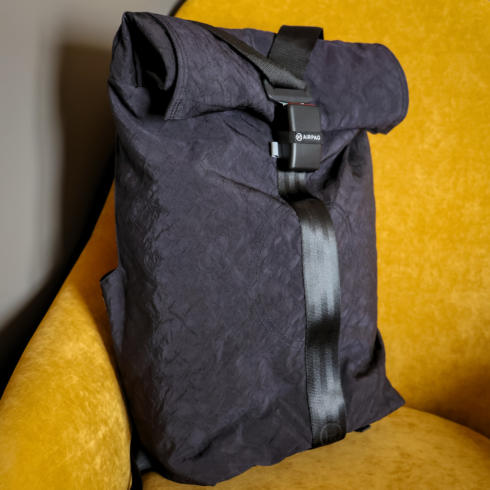 Schwarzer Rolltop-Rucksack mit schwarzem Riemenverschluss auf einem senfgelben Sessel.