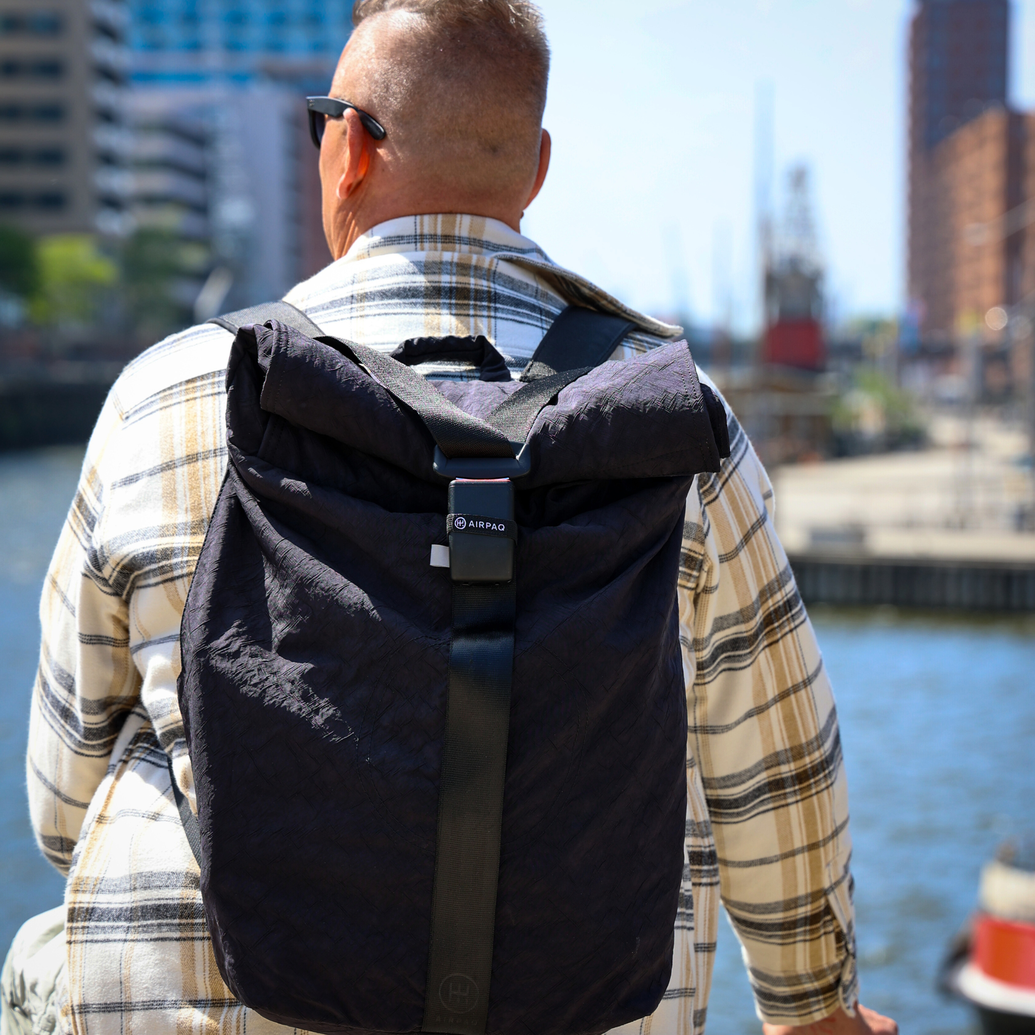 Person mit kariertem Hemd und schwarzem Rucksack steht am Wasser; im Hintergrund sind urbane Gebäude zu sehen.