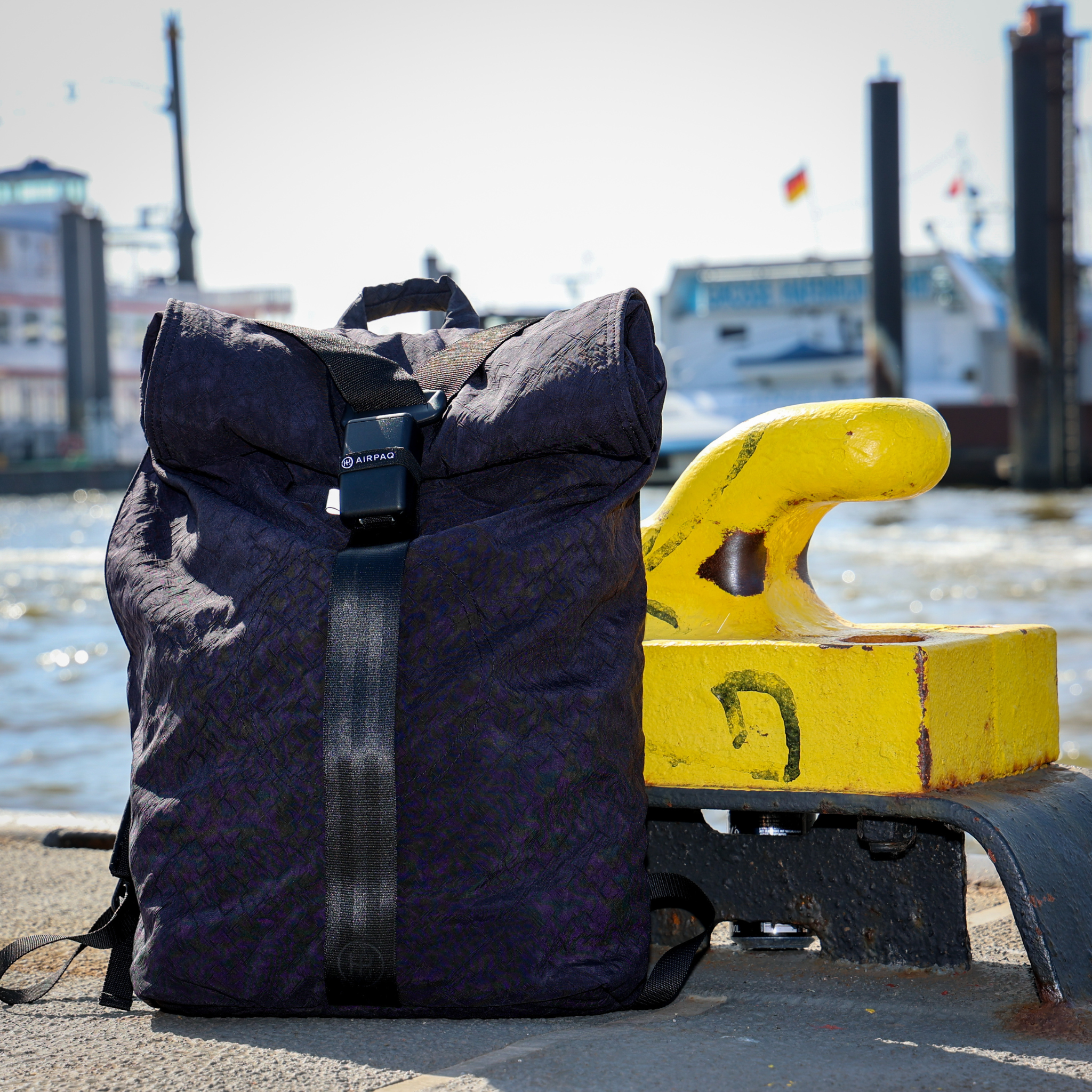 Lila Rucksack neben einer gelben Ankerwinde auf einem Hafendock, im Hintergrund ein unscharfes Schiff auf dem Wasser.