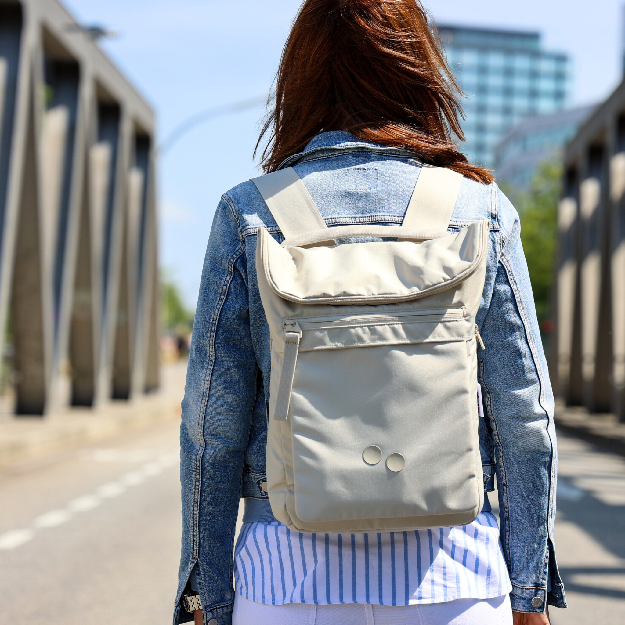 Frau mit beigefarbenem Rucksack und Jeansjacke geht über Brücke in urbaner Umgebung.