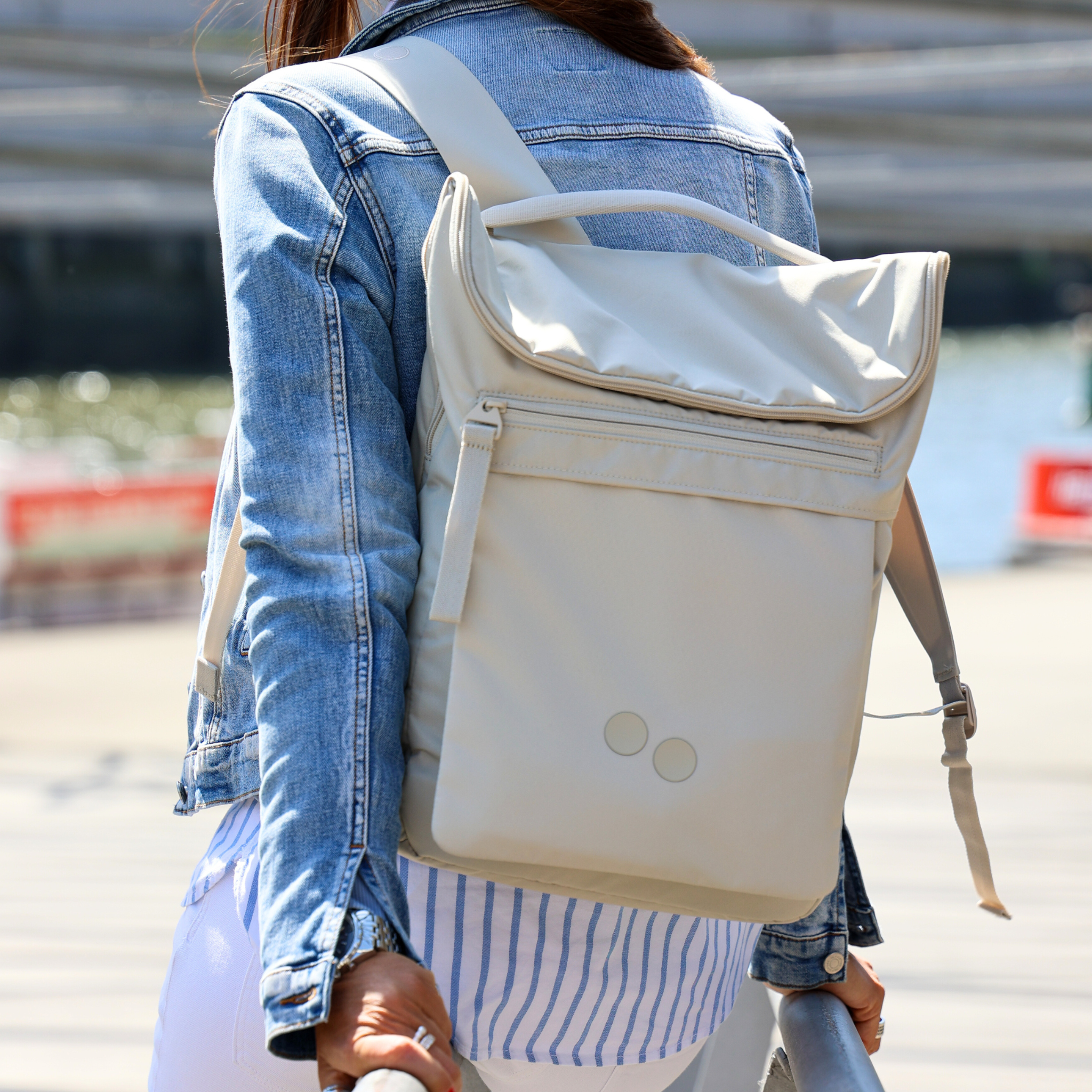 Eine Person mit Jeansjacke trägt einen cremefarbenen Rucksack und spaziert an einem sonnigen Tag entlang einer Uferpromenade.