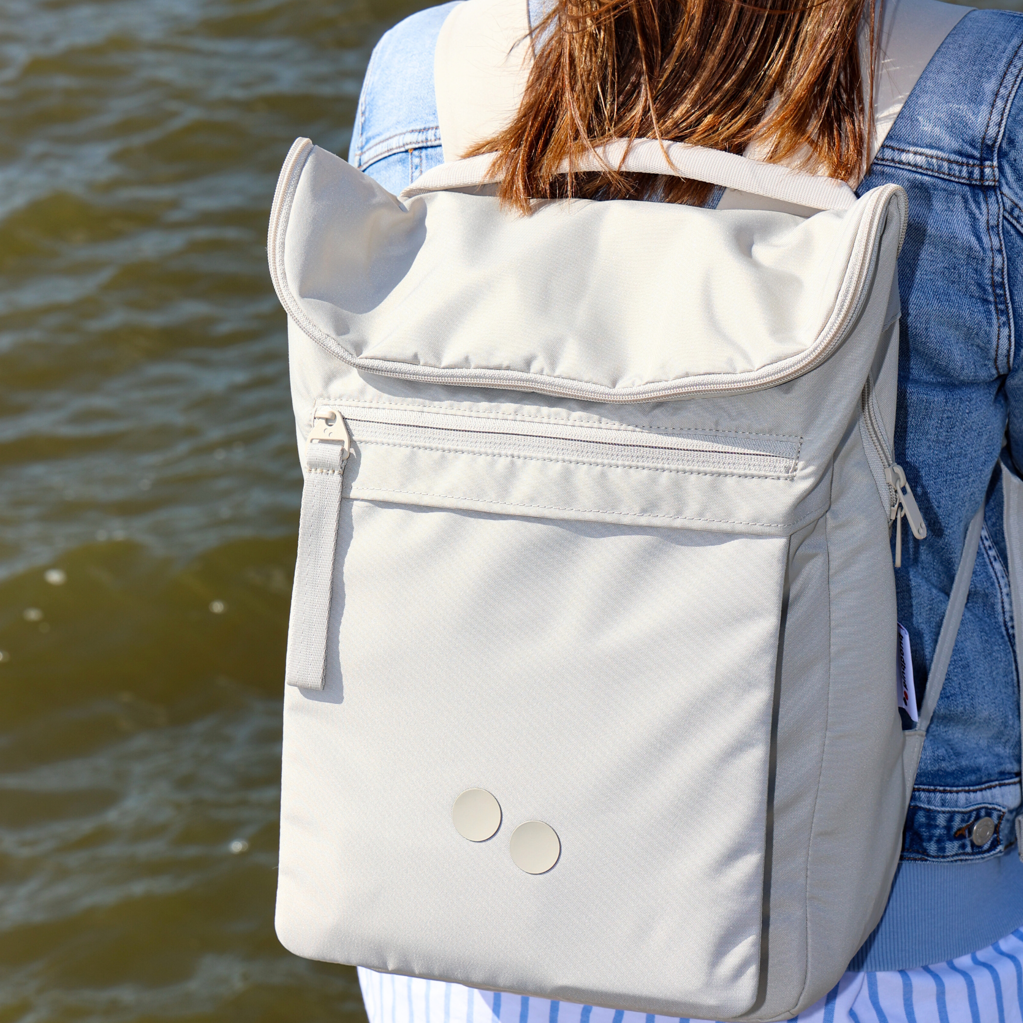 Person mit beigefarbenem Rucksack und Jeansjacke steht vor einem Gewässer.