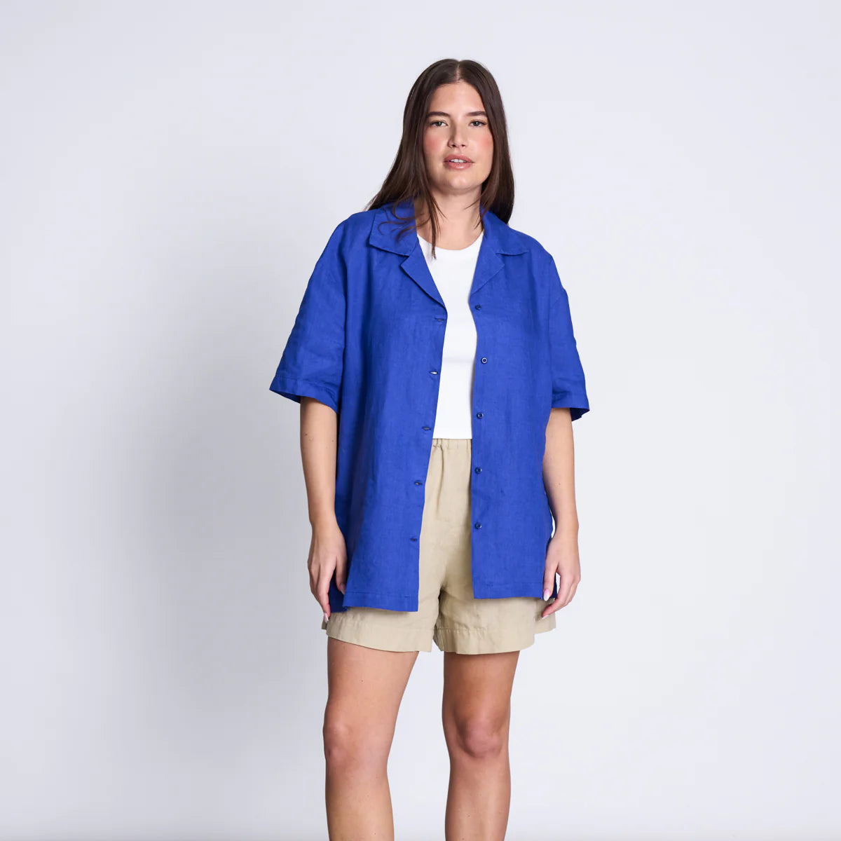Frau in weißem T-Shirt und blauer, offener Bluse, trägt beige Shorts, steht vor einem schlichten, grauen Hintergrund.