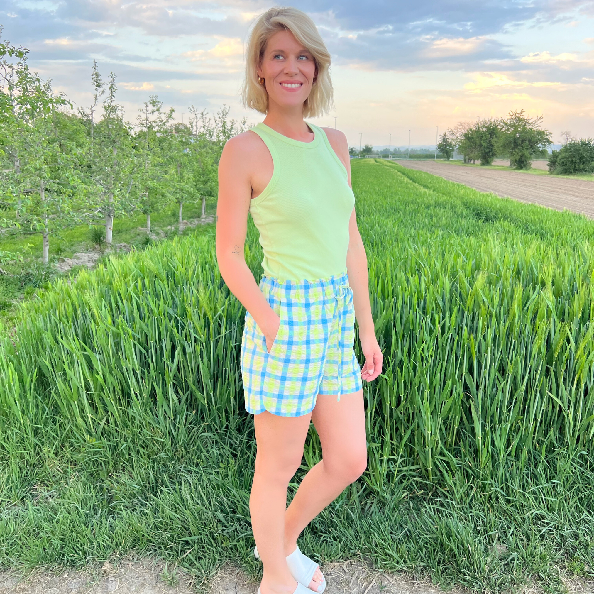 Frau in grünem Top und karierten Shorts steht auf einem Feldweg neben grünem Feld und Bäumen bei Sonnenuntergang.