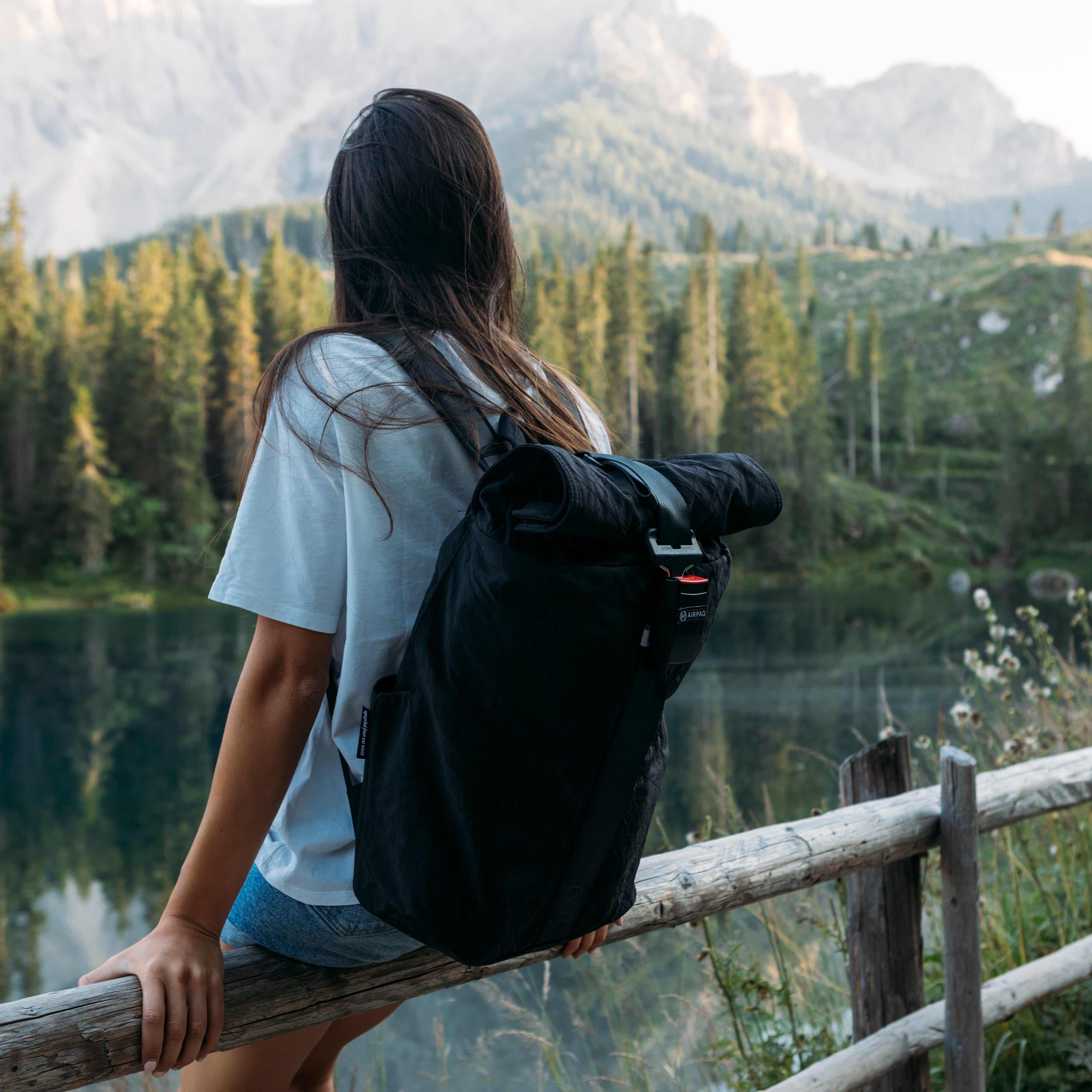 Eine Person mit langen Haaren und einem schwarzen Rucksack sitzt auf einem Holzzaun und blickt an einem klaren Tag auf einen bewaldeten See mit Bergen im Hintergrund.