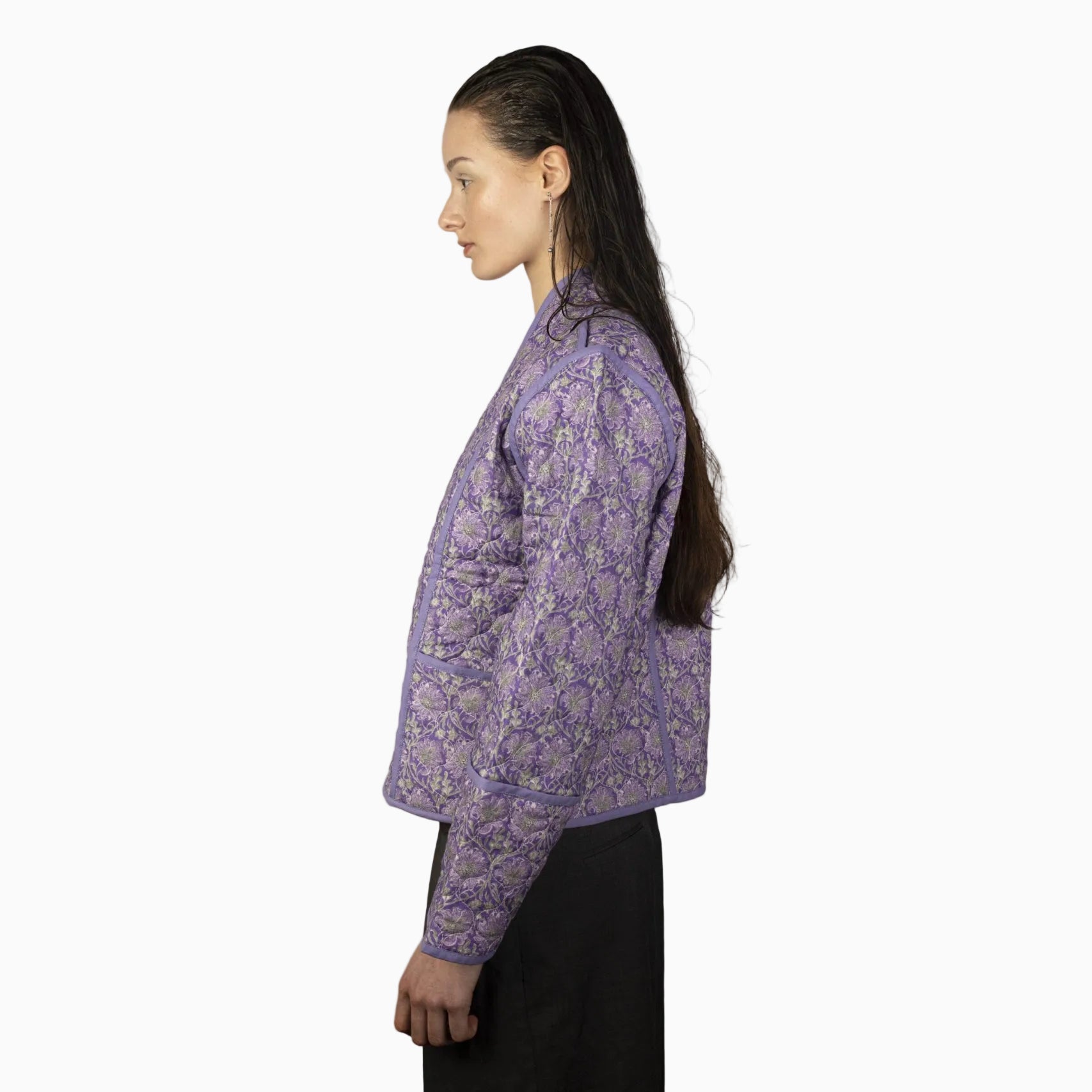 Eine Frau mit langem, nassem, zurückgekämmtem Haar steht im Profil vor einem schlichten Hintergrund und trägt eine lavendelfarbene Steppjacke mit Blumenmuster und eine dunkle Hose.