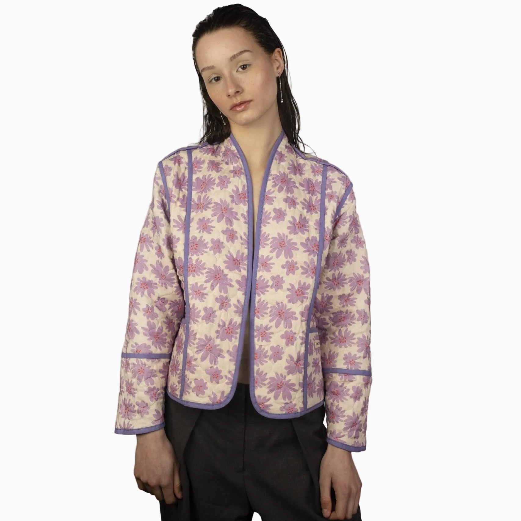 Eine Person mit glattem, feucht wirkendem Haar trägt eine helle Steppjacke mit rosa und lila Blumenmuster und lavendelfarbenem Besatz über einer grauen Hose vor einem schlichten, hellen Hintergrund.