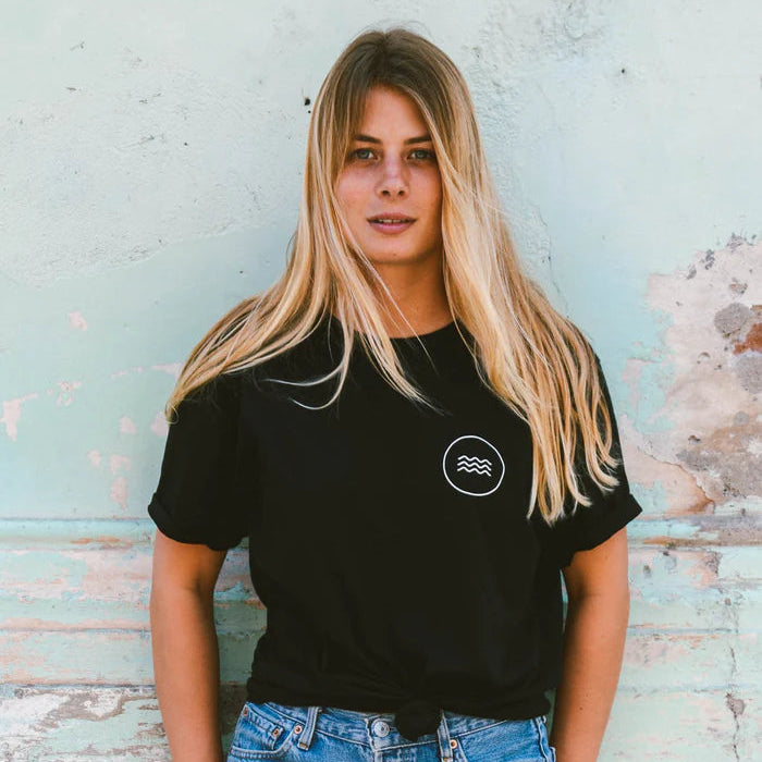 Eine junge Frau mit langen blonden Haaren steht vor einer verwitterten, blassblauen Wand. Sie trägt ein schwarzes T-Shirt mit einem kleinen weißen Wellenlogo und ausgefranste Jeansshorts und schaut selbstbewusst in die Kamera.