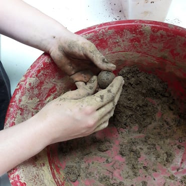 Die Hände einer Person formen eine kleine Kugel aus feuchter Erde oder Ton in einer großen, roten Schale, die mit loser Erde oder Ton gefüllt ist.