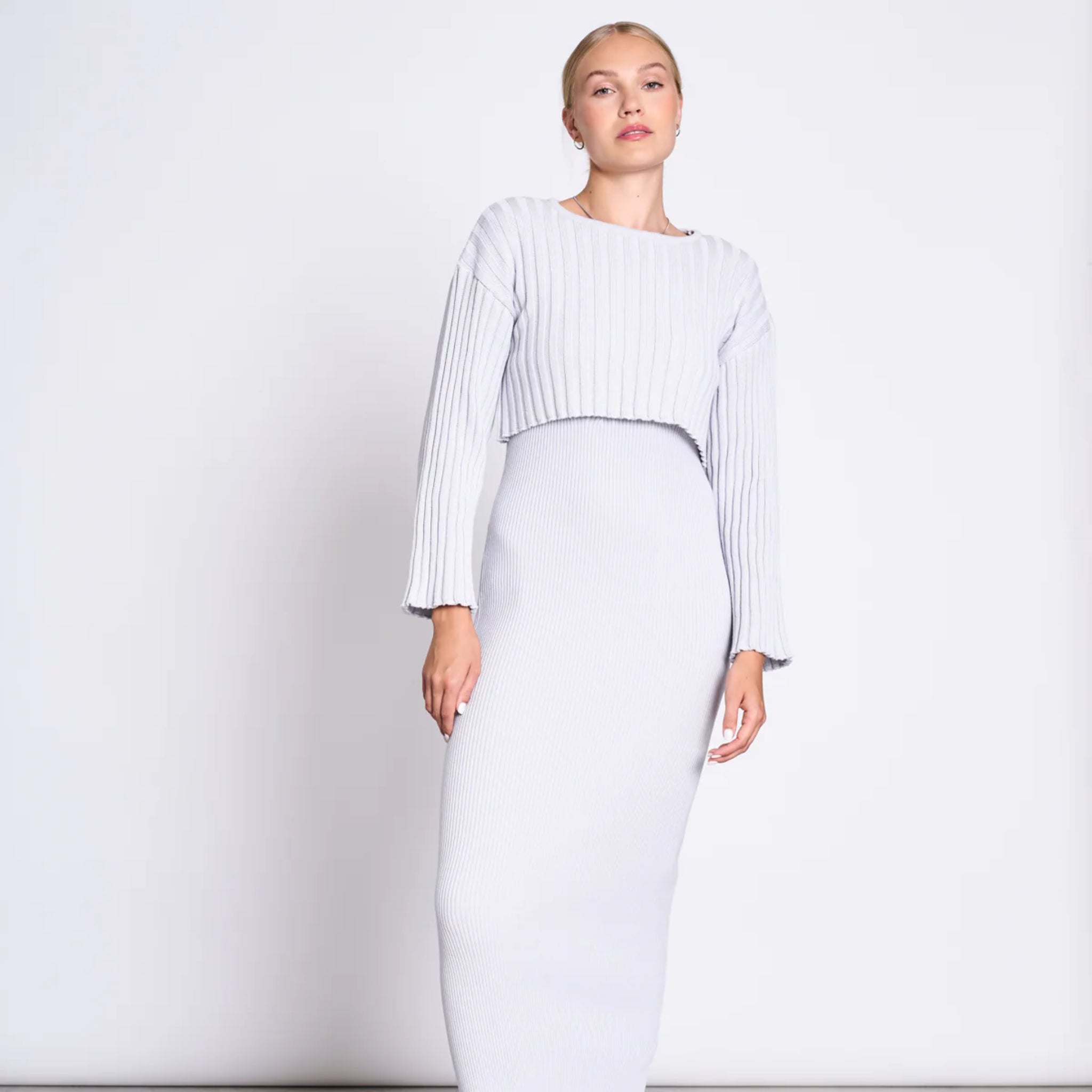 Person in einem eleganten, hellgrauen Strickoutfit mit langem Rock und langärmligem Top vor neutralem Hintergrund.