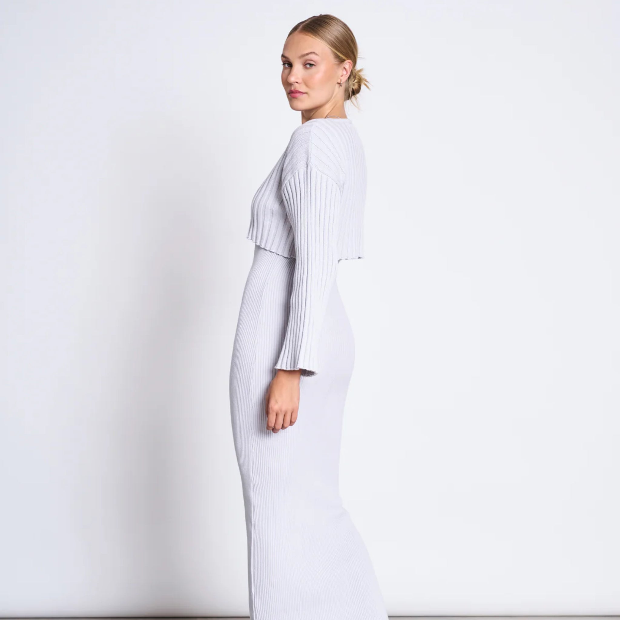 Frau in grauem, langärmligem, geripptem Oberteil und passendem langen Rock posiert seitlich vor weißem Hintergrund.