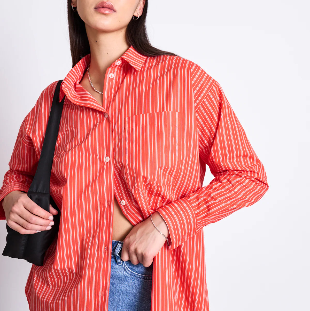 Eine Frau, die ein rot-weiß gestreiftes Button-up-Hemd und blaue Jeans trägt, hält mit einer Hand einen Teil ihres Hemdes offen und trägt eine schwarze Tasche über ihrer Schulter. Sie hat langes dunkles Haar und steht vor einem schlichten weißen Hintergrund.